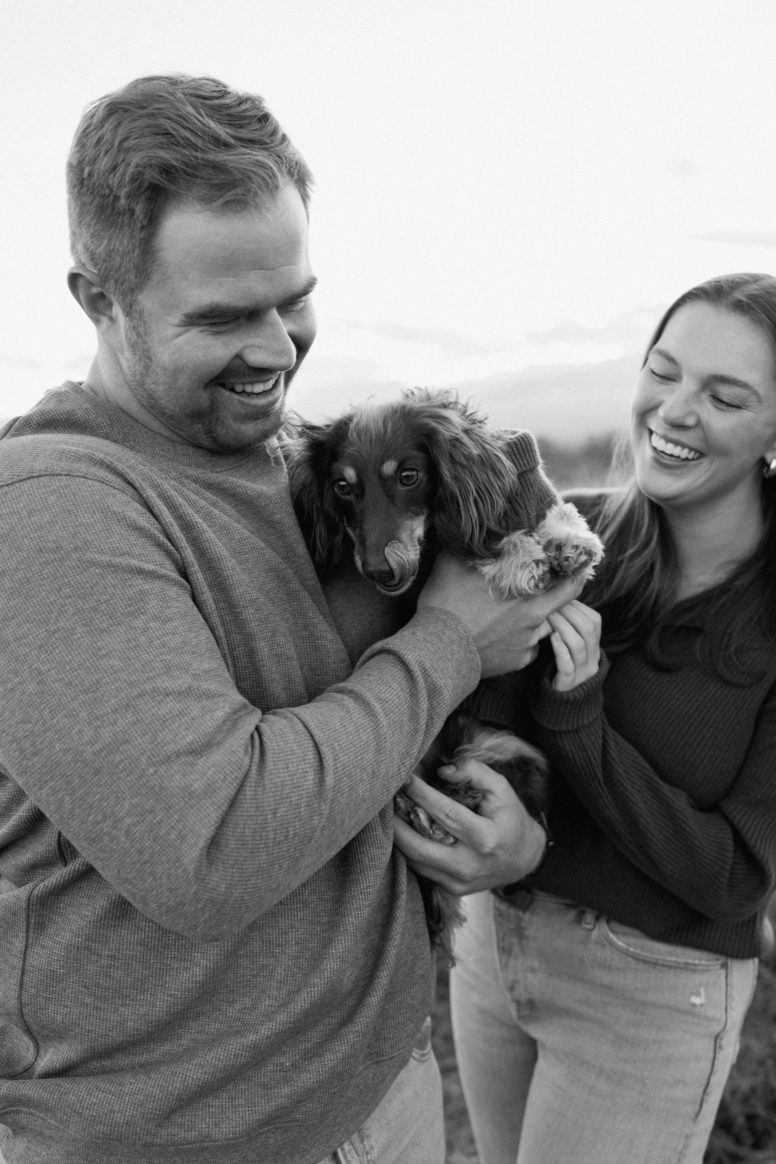 A happy couple holding a small dog outdoors, smiling and laughing in black and white.