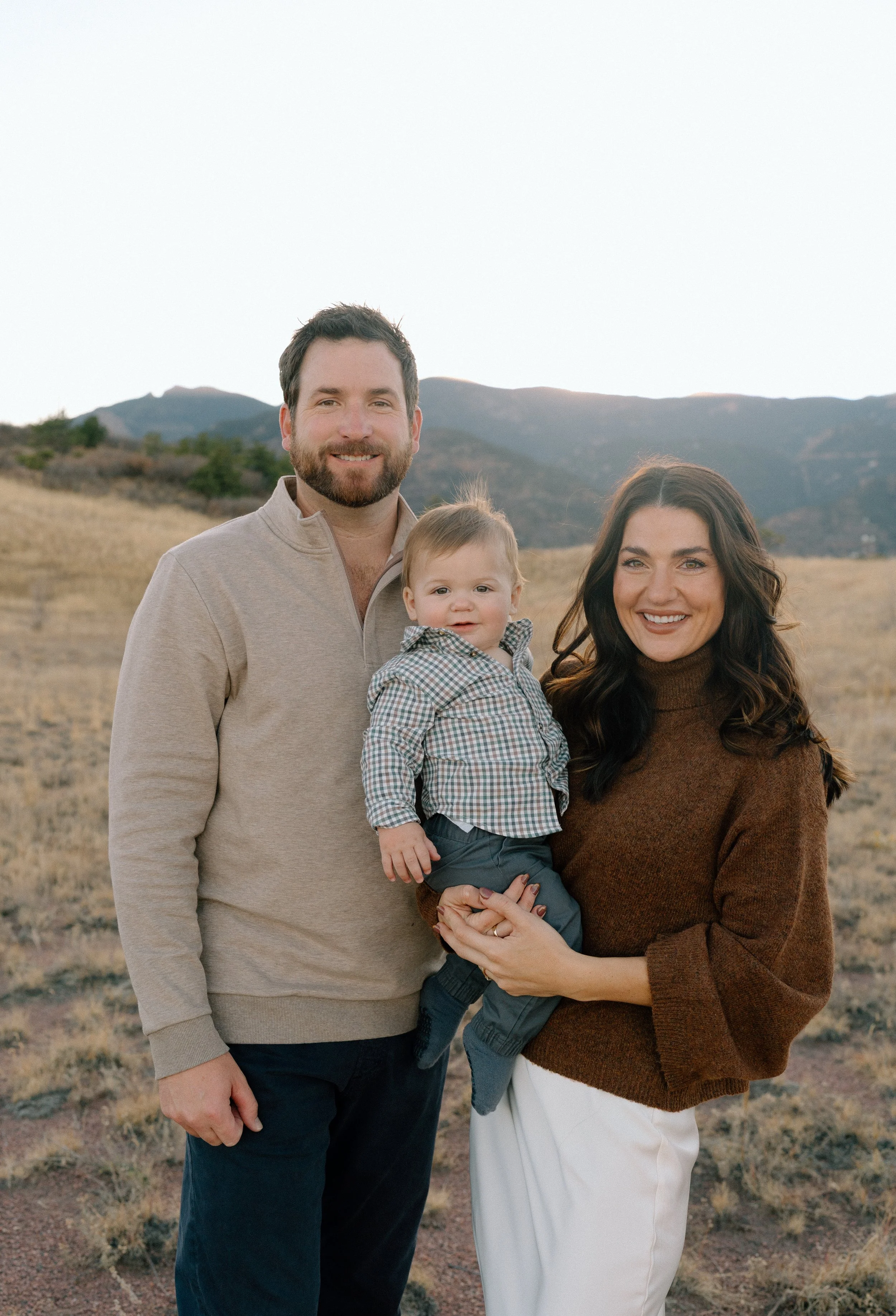 A family of three standing outdoors on a grassy field with mountains in the background, smiling at the camera. The man has a beard, wearing a light beige sweater and dark pants. The woman has dark wavy hair, wearing a brown sweater and white pants, h