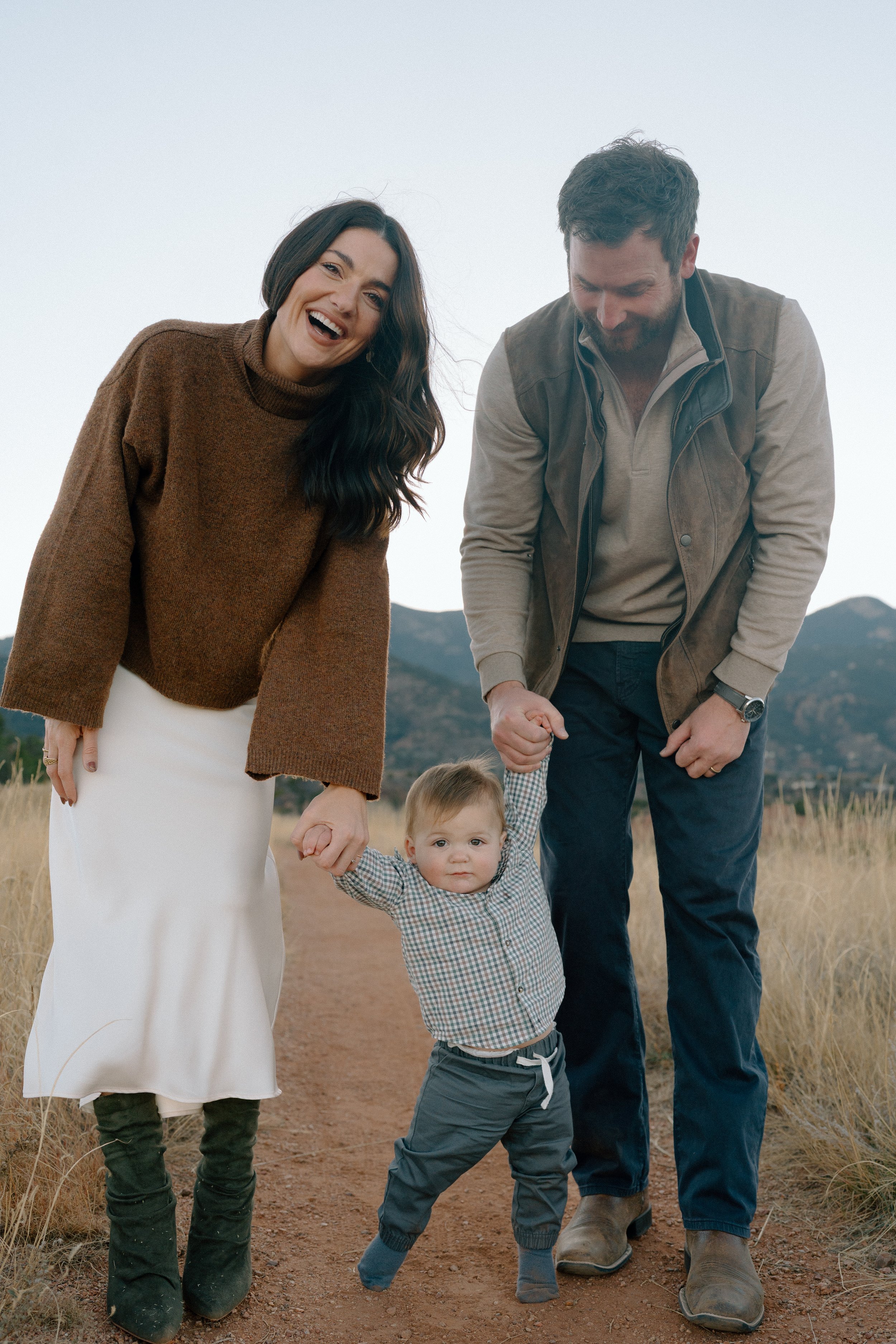 A family of three walking outdoors on a dirt path in the mountains, with the parents holding their child's hands and smiling. The mother has dark hair and wears a brown sweater and white skirt. The father has a beard and wears a beige shirt under a b
