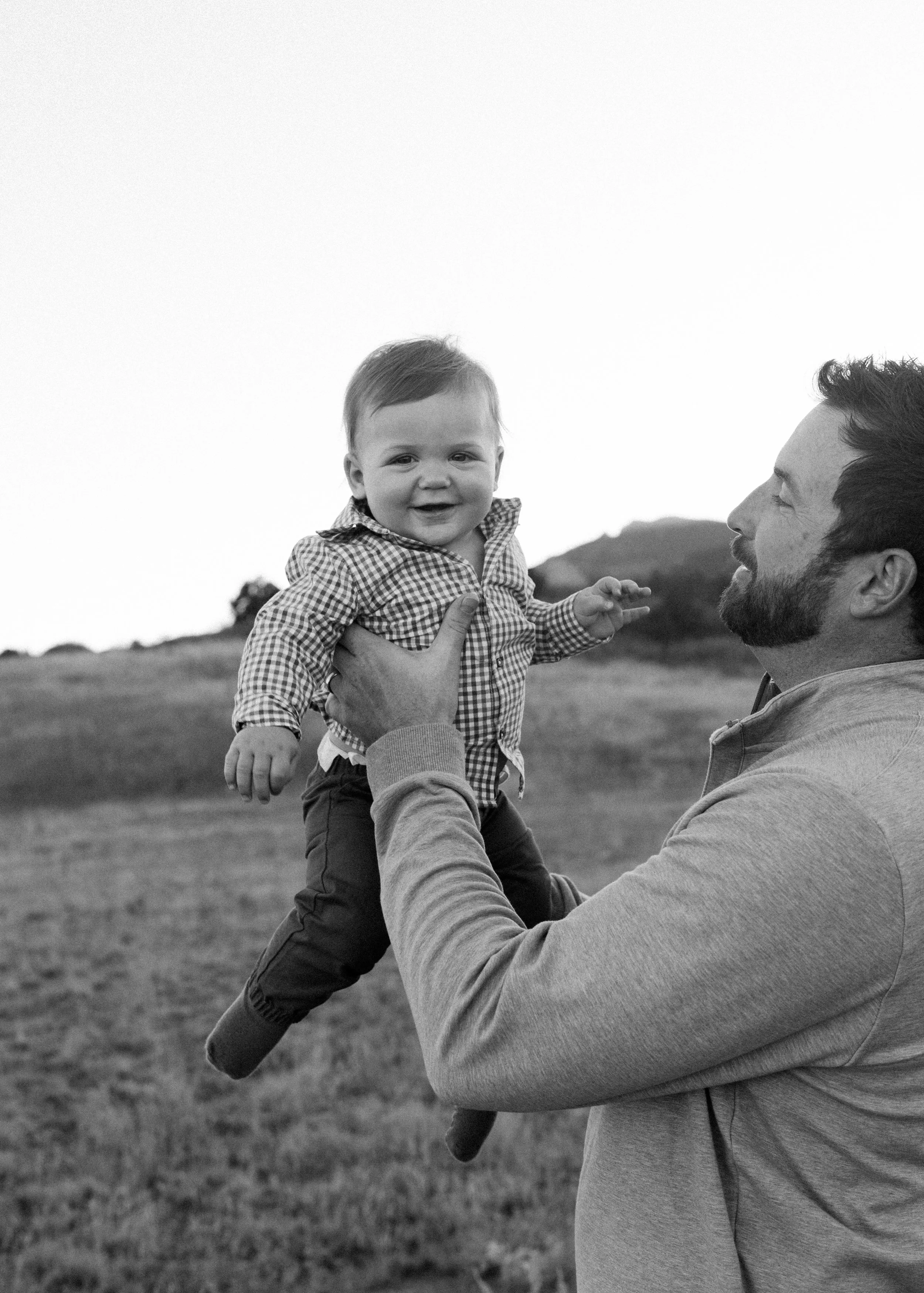 A man lifting a smiling baby outdoors in a field.
