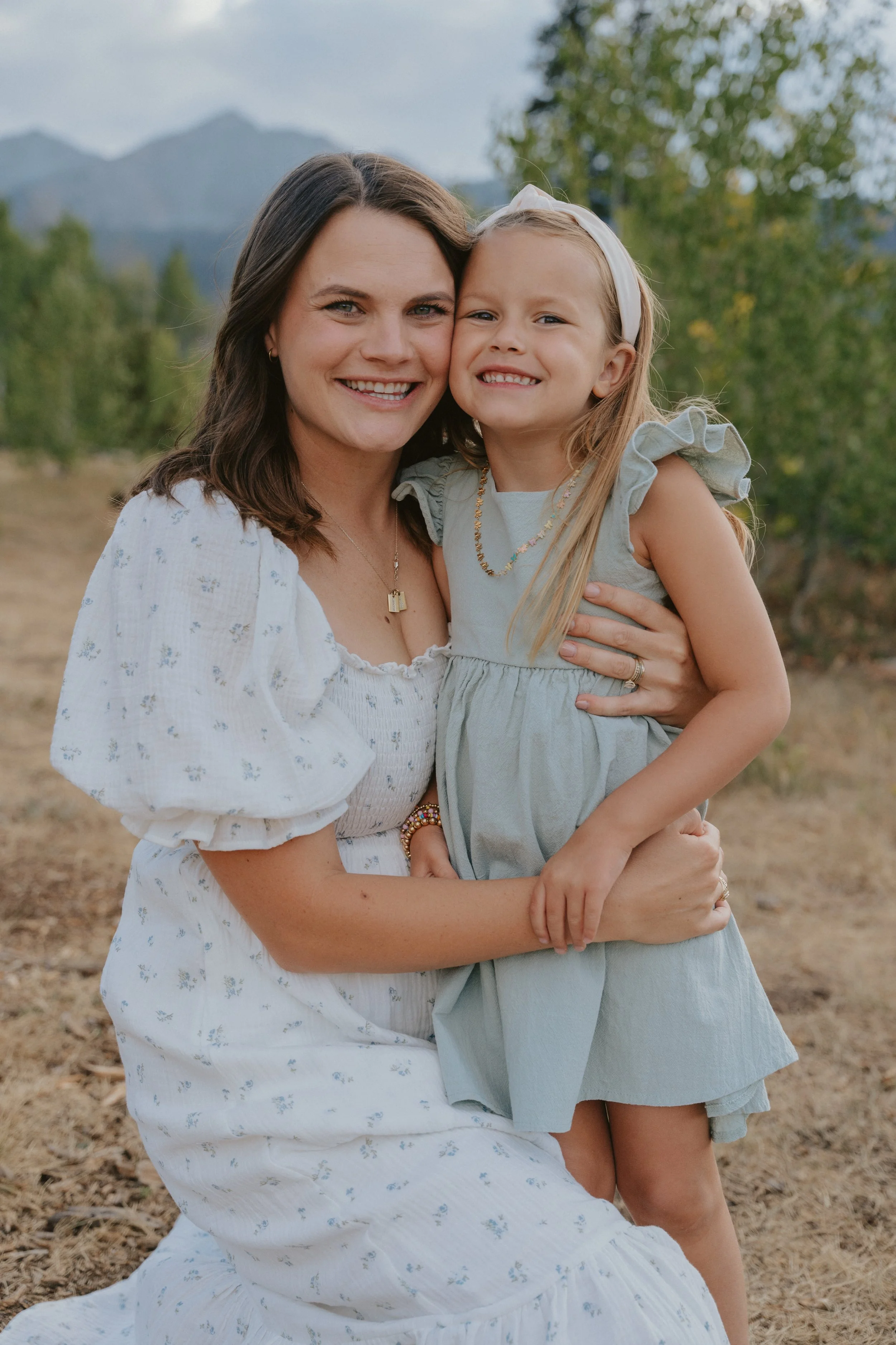 A woman and a young girl smiling and hugging outdoors with mountains and trees in the background.