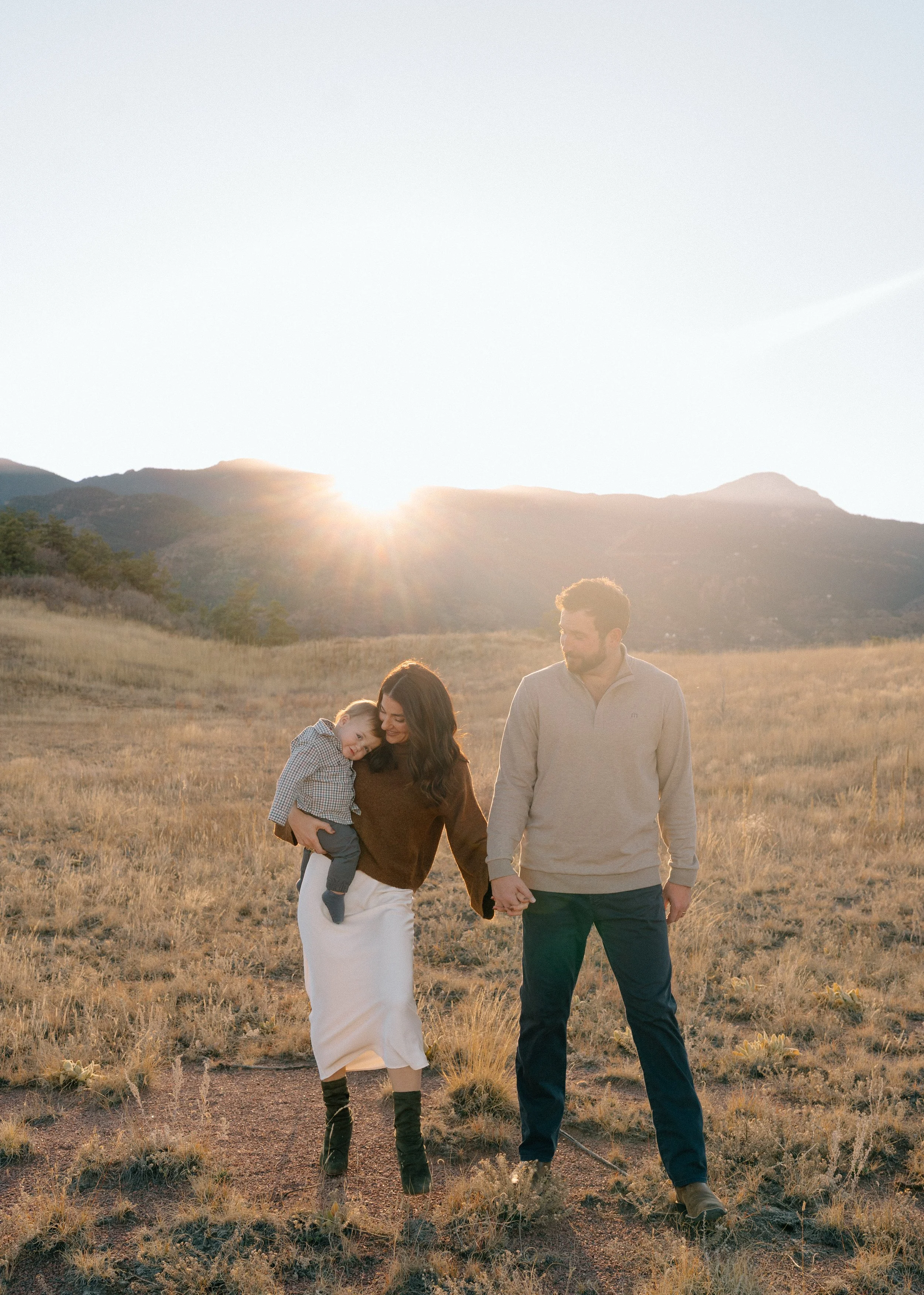 A family of three walking hand-in-hand through a grassy field at sunset, with mountains in the background.