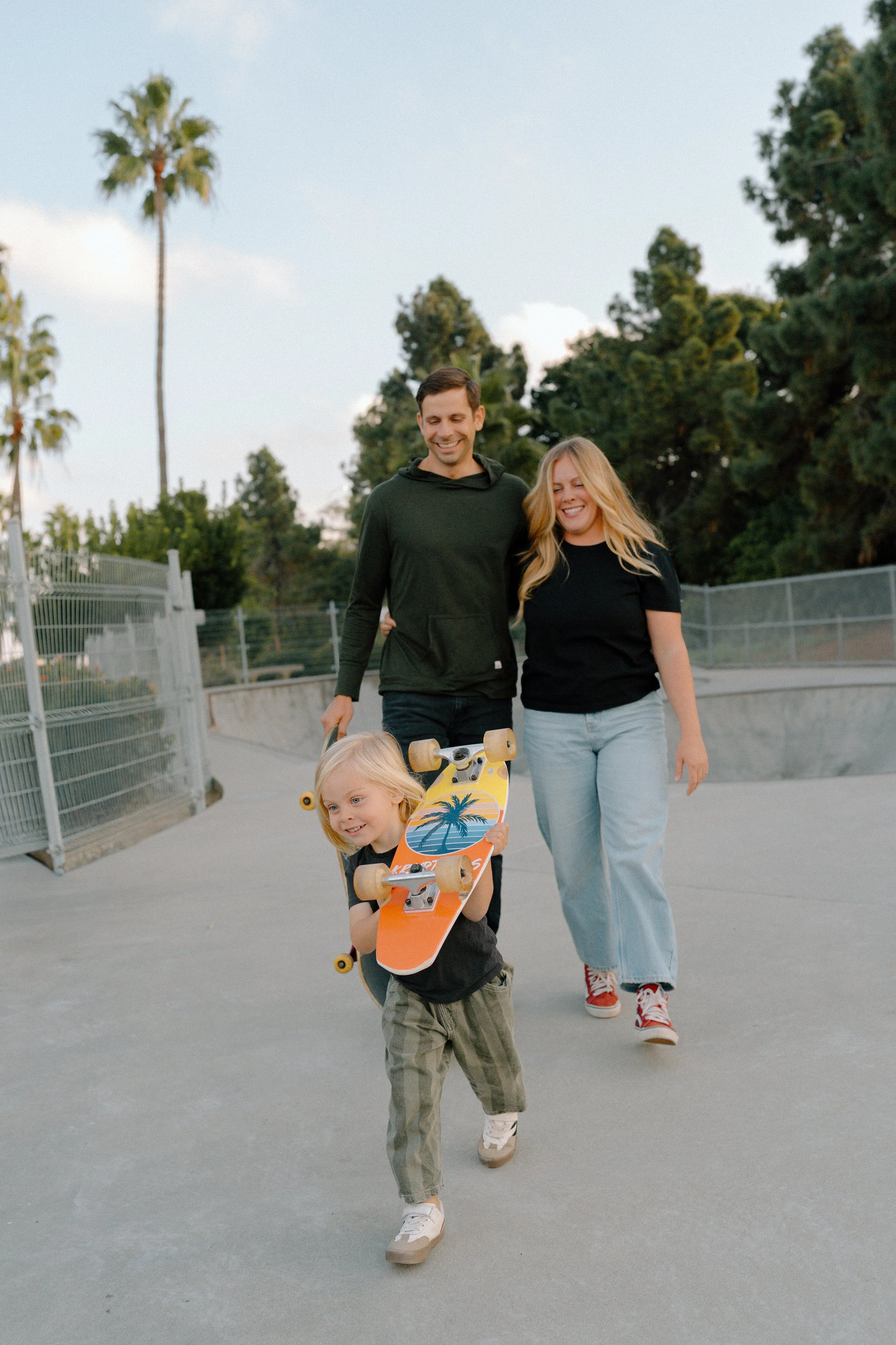 A family of three, with a young girl holding a skateboard, walking at a skate park during late afternoon or early evening. The father is smiling and wearing a dark hoodie, the mother is smiling and wearing a black t-shirt and light jeans. The skate p