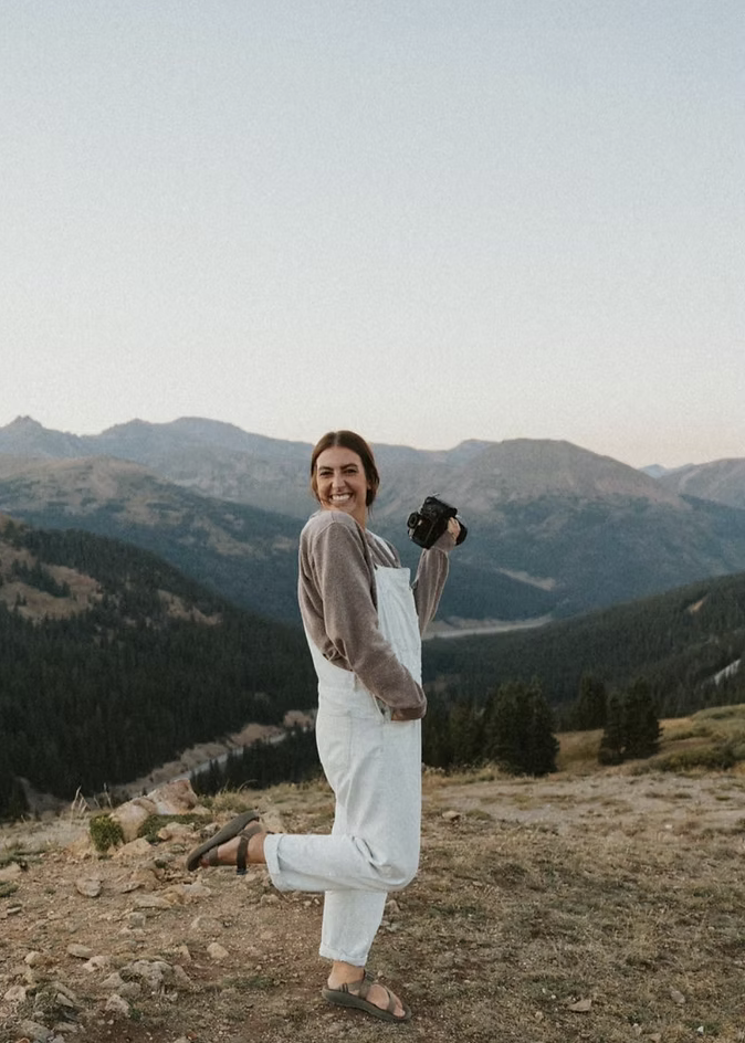 A woman smiling and holding a camera stands outdoors on a mountain landscape during daylight, wearing a gray hoodie, white overalls, and sandals.