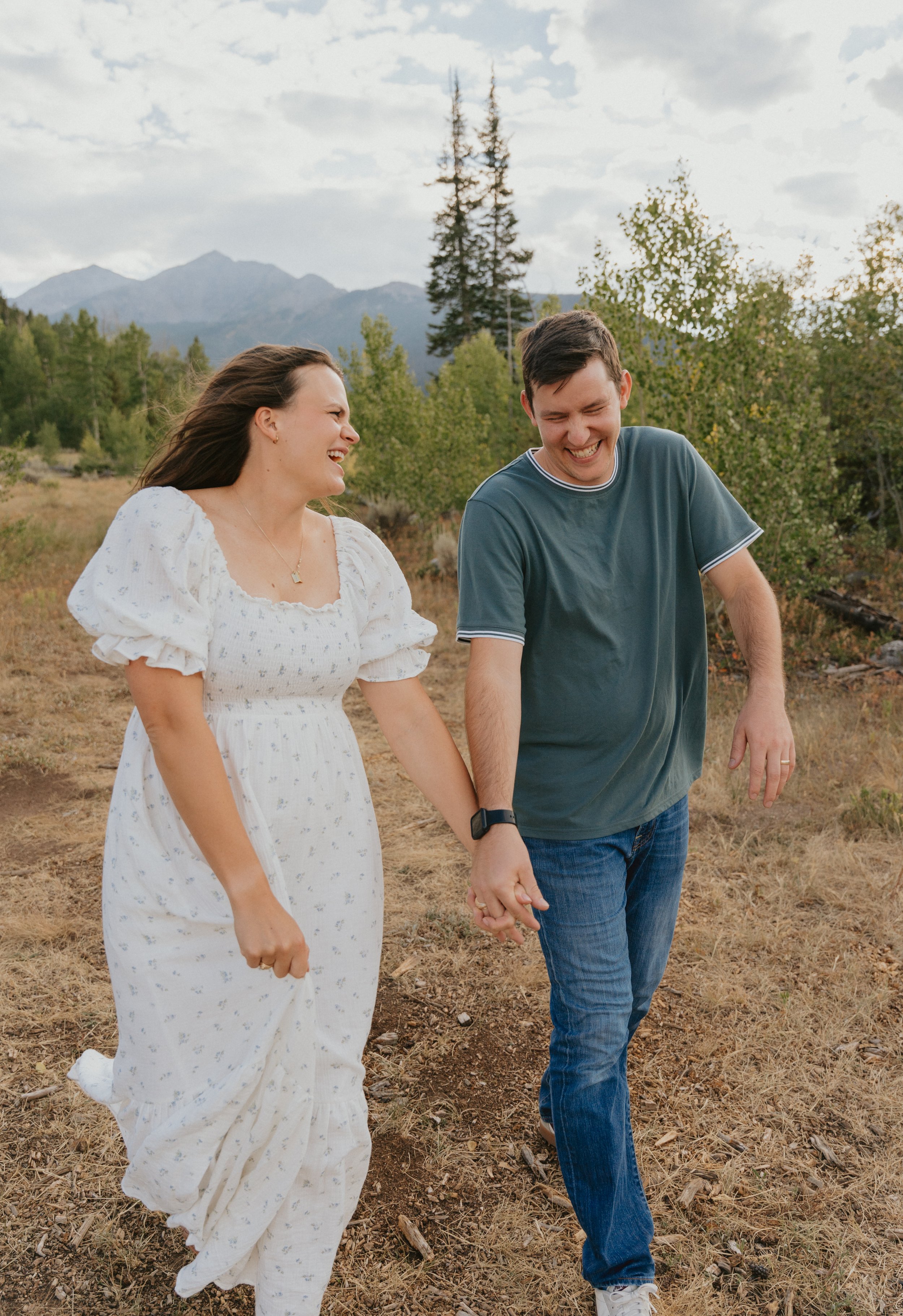 A couple walking hand-in-hand outdoors, smiling and laughing, in a natural setting with mountains, trees, and cloudy sky in the background.