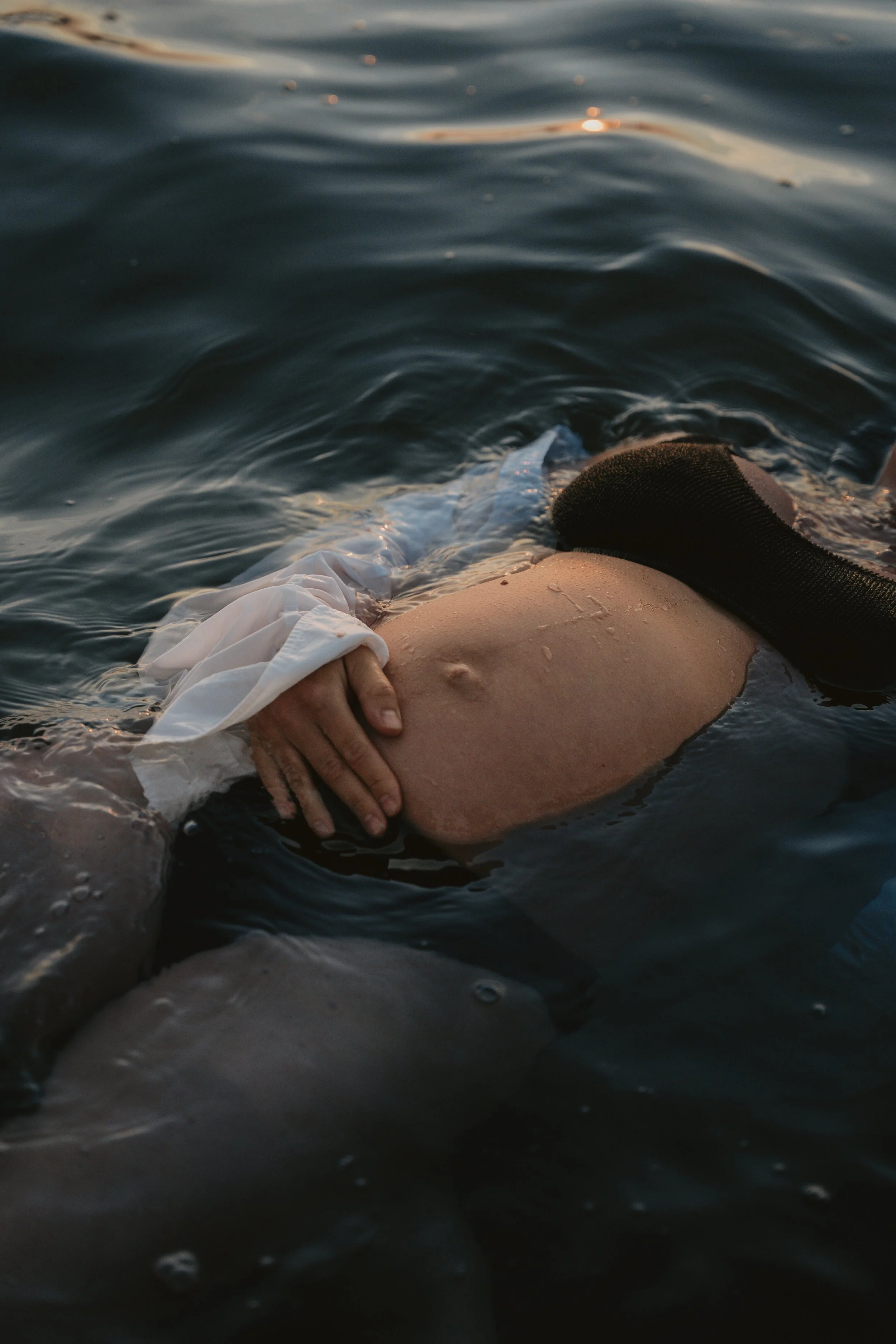 A person in a white shirt holding their pregnant belly while floating in dark water, with a black strap across the belly and a grayish object partially submerged nearby.
