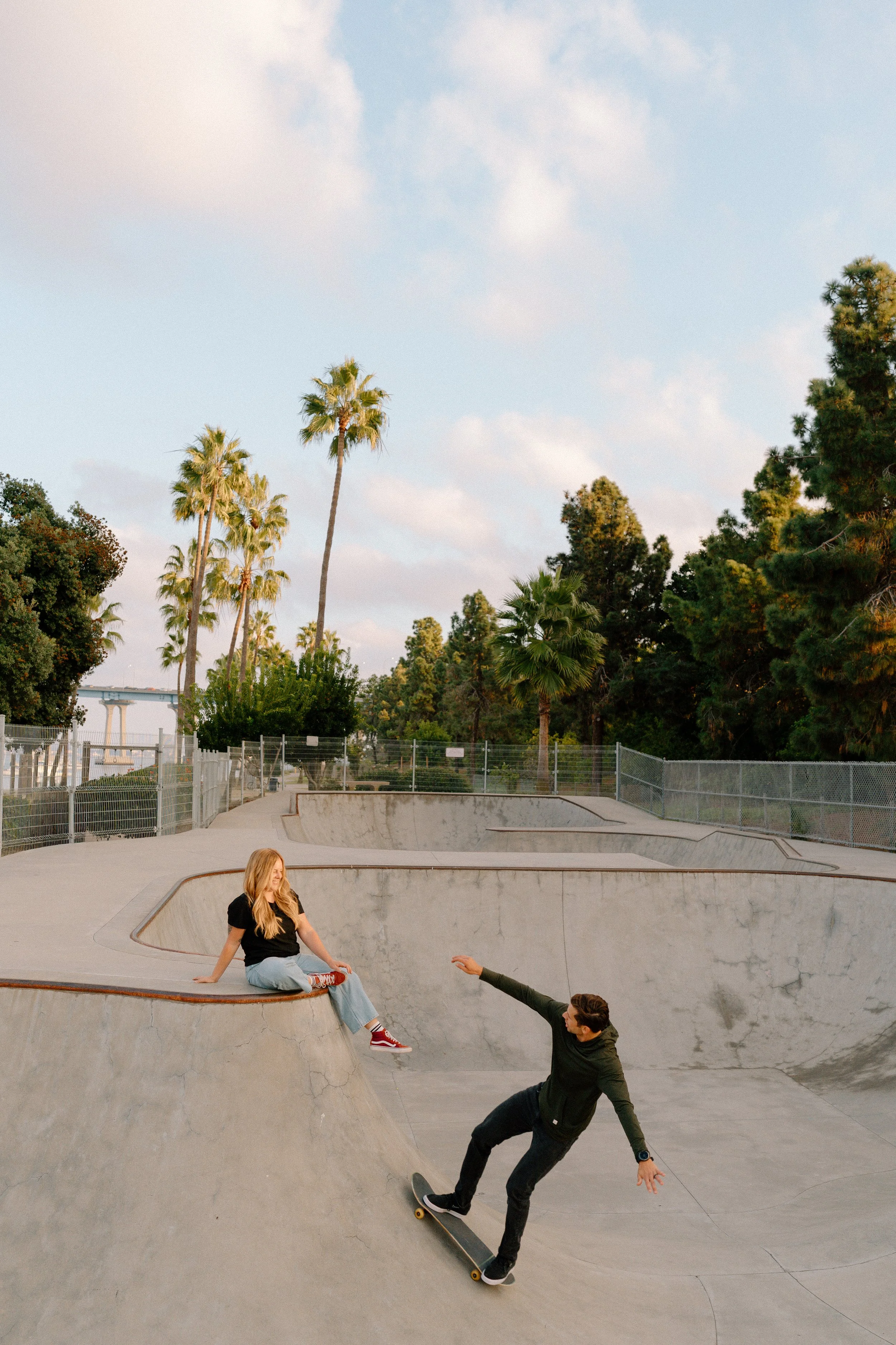 A young man skateboarding at a concrete skate park with a young woman sitting on the edge, with palm trees and a partly cloudy sky in the background.