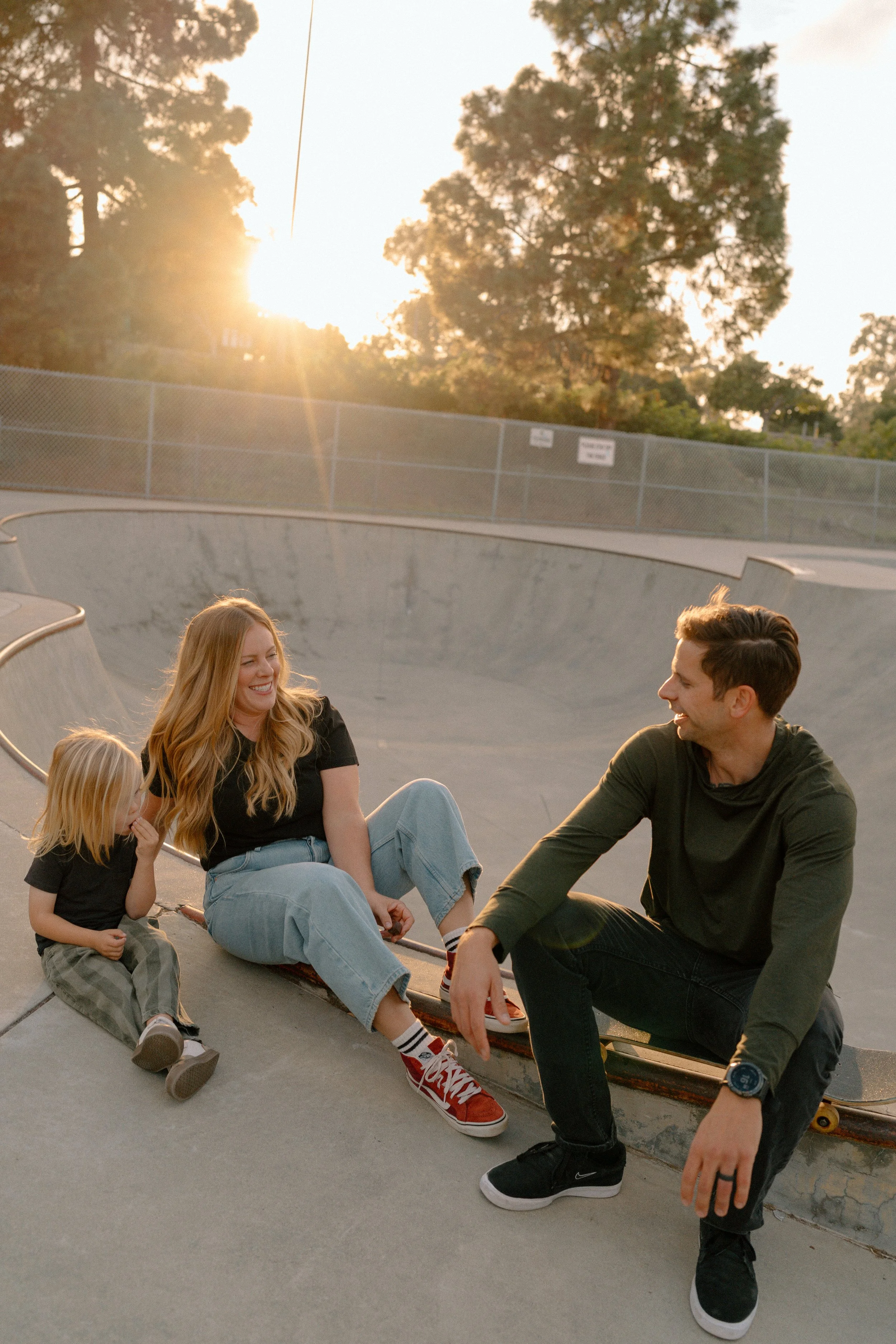 Three people sitting and laughing at a skate park, with a concrete bowl and trees in the background during sunset.