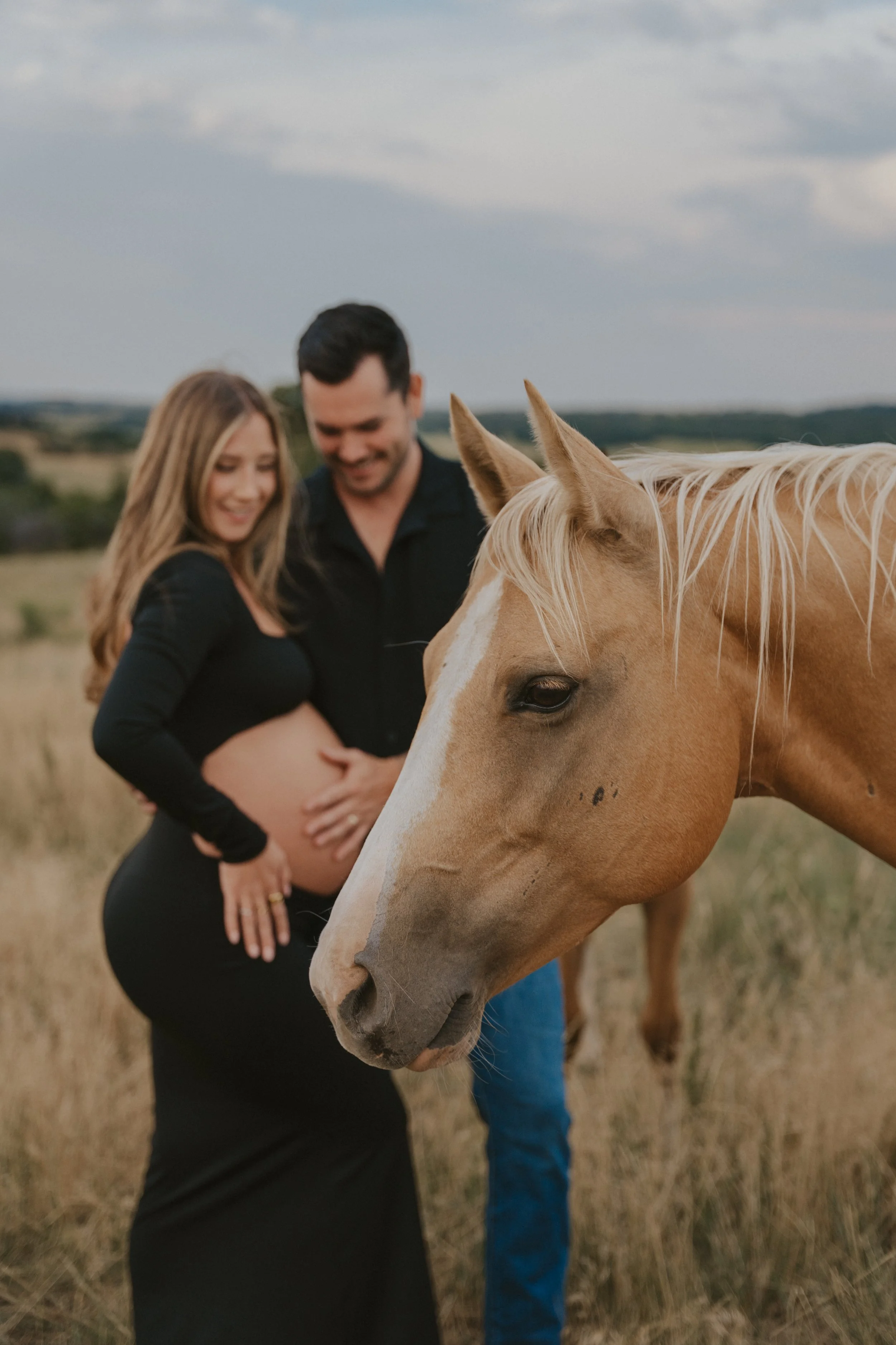 Pregnant woman and man smiling with a horse in an outdoor field.