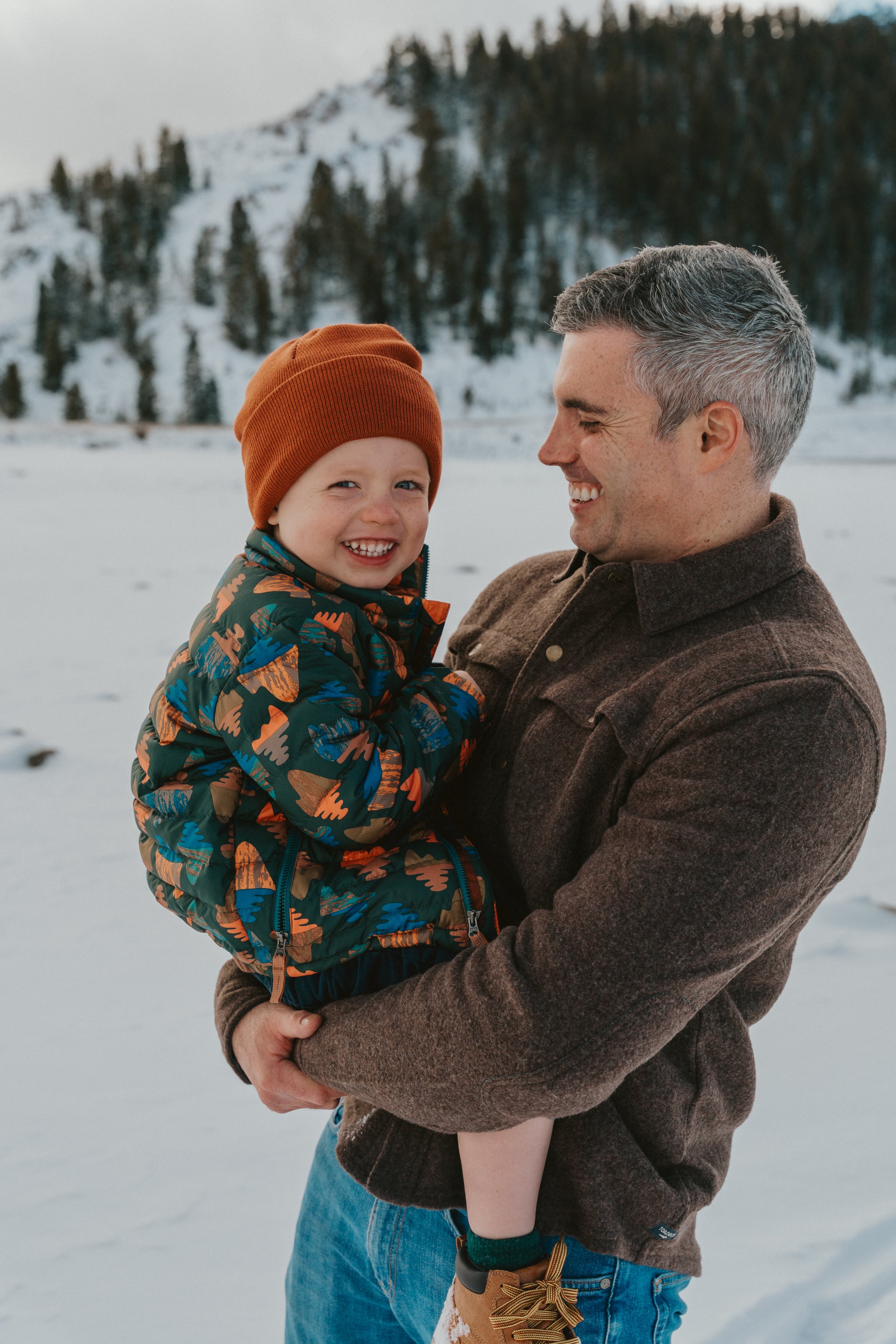 A man holding a smiling young boy in a snowy outdoor setting with mountains and trees in the background.