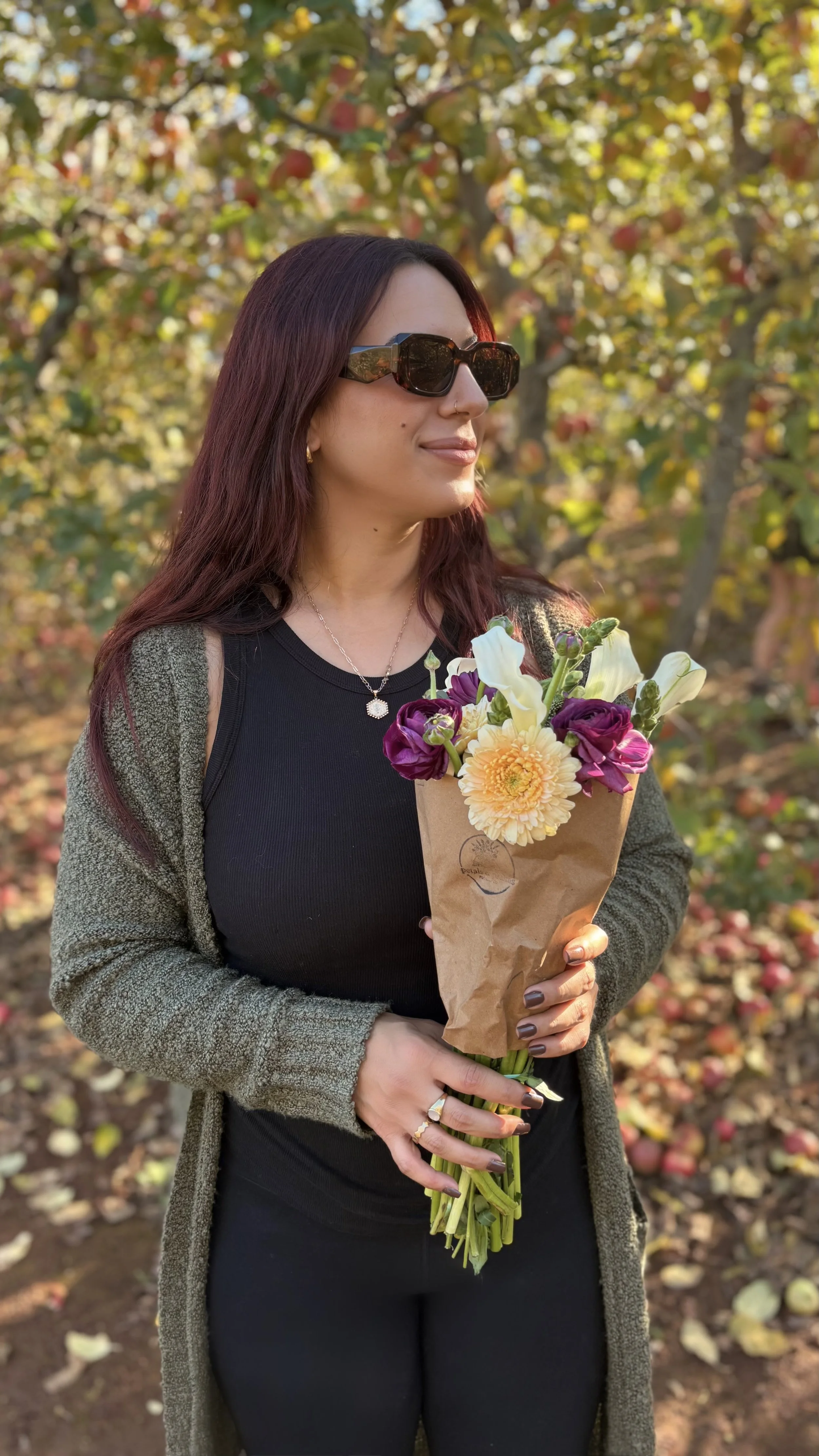 A woman with long reddish-brown hair, wearing sunglasses, a black top, a gray cardigan, and a necklace, holding a bouquet of flowers in a paper wrapping, standing outdoors among trees with autumn leaves.