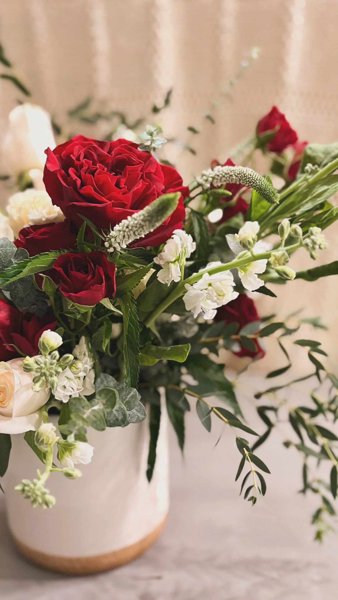 A floral arrangement in a cream-colored vase with a light brown base, featuring red roses, white flowers, green leaves, and baby’s breath.