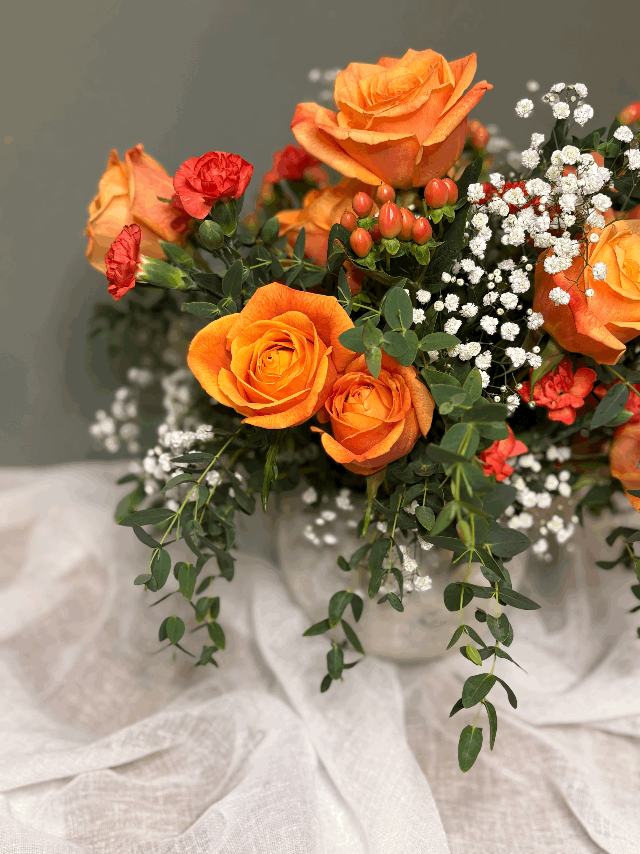 A bouquet of orange and red roses with white baby's breath and green foliage, placed on a white fabric surface.