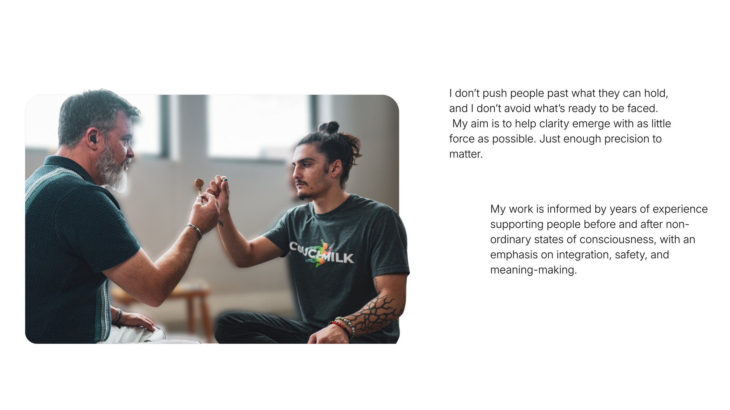 A young man with long dark hair in a bun and patterned tattoos on his arm, wearing a black t-shirt, practices finger painting on the finger of an older man with gray hair and beard, inside a bright room with large windows.