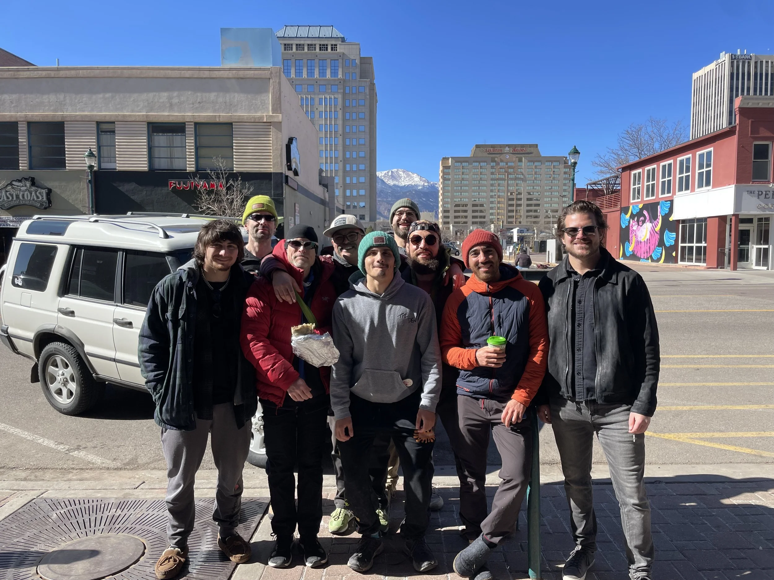 Group of nine people standing on sidewalk in downtown area with buildings and mountains in background.