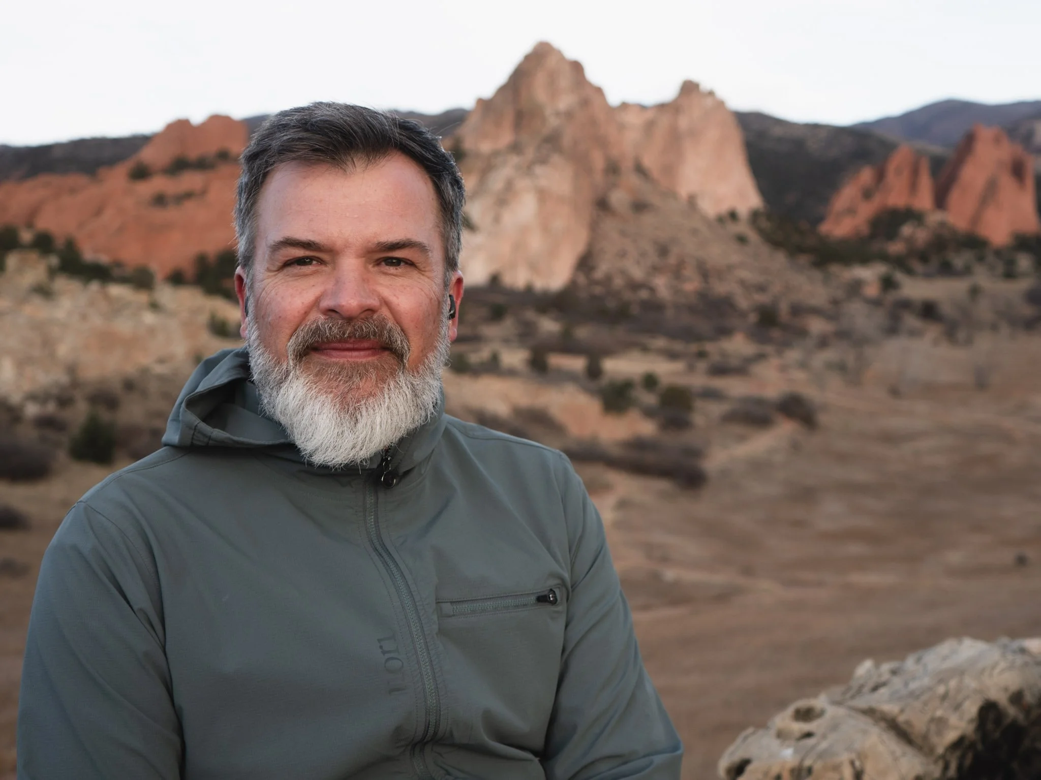 A middle-aged man with a beard and mustache, wearing a gray jacket, outdoors with rocky desert landscape and large red rock formations in the background.