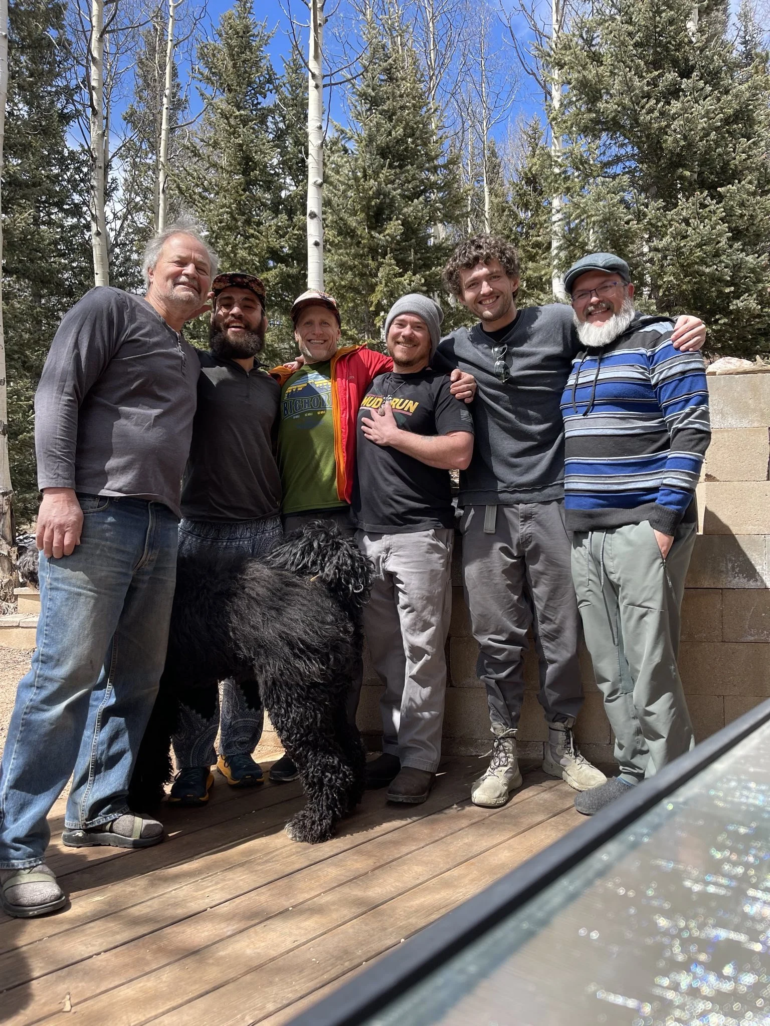 Seven men standing outdoors on a wooden deck, some with arms around each other, with a large black dog in front of them. They are surrounded by tall evergreen and aspen trees under a bright blue sky.