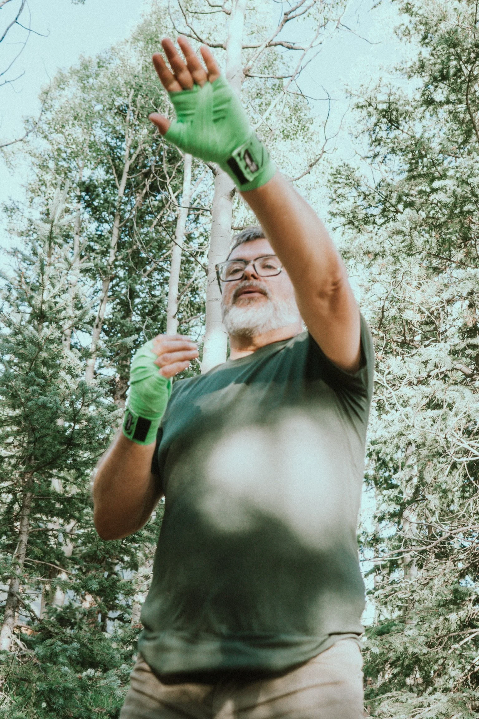An older man with glasses and a beard standing outdoors among trees, wearing green gloves and a dark t-shirt, reaching out with his right hand.