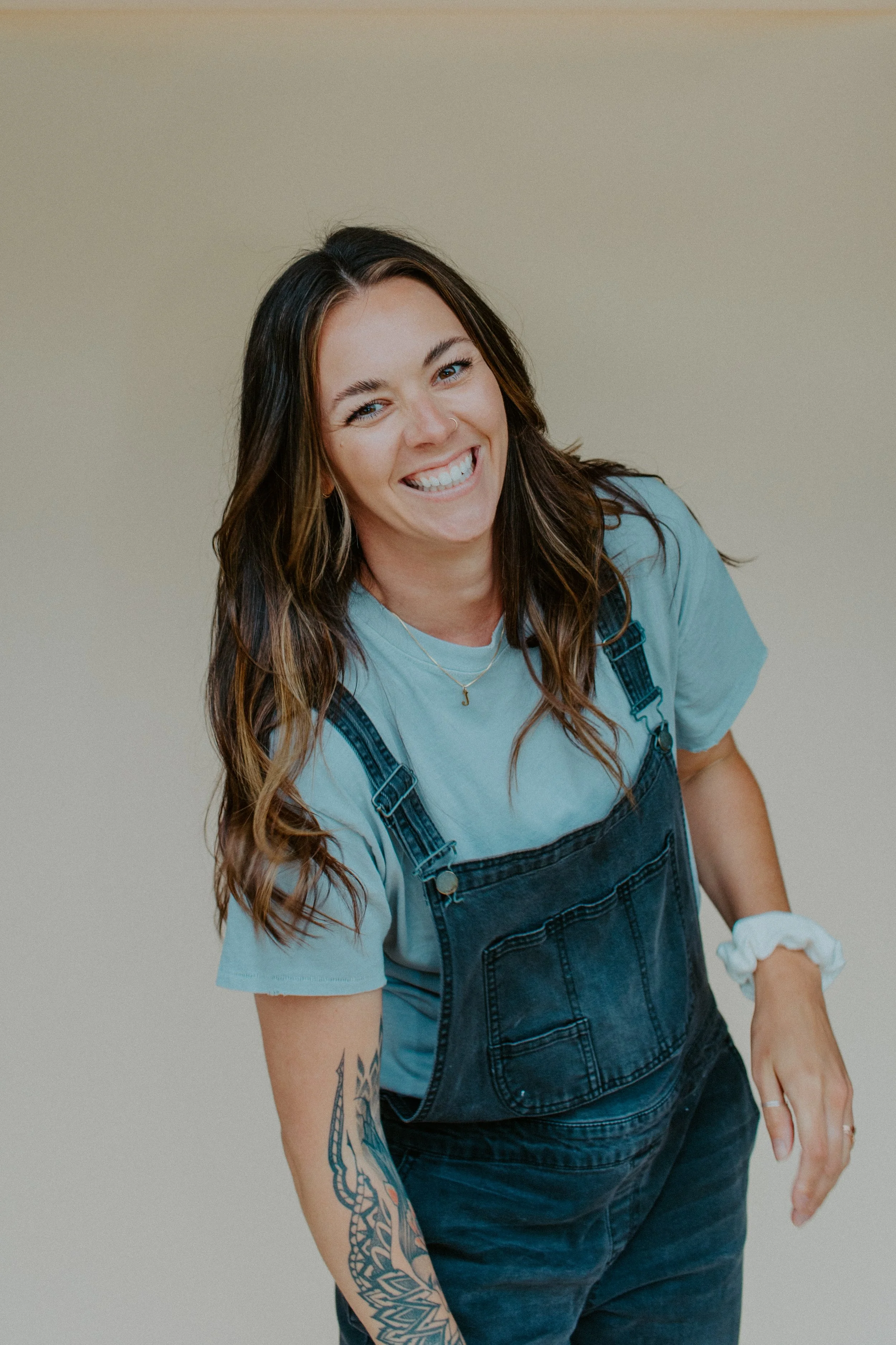 Woman with long brown hair smiling, wearing a light blue t-shirt, dark denim overalls, with tattooed arm, leaning toward the camera against a plain beige background.