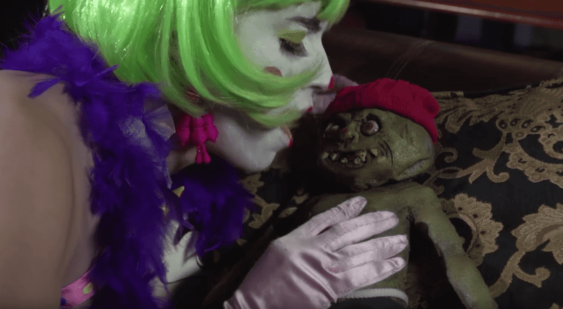 A person with a green wig, clown makeup, and purple feathered costume holds a creepy doll with a red hat and frightening face.