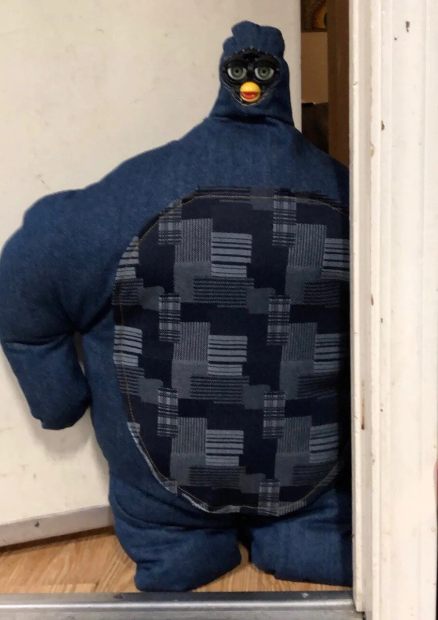 A person wearing a hoodie with a chicken mask, which has glasses and a yellow beak, standing near a door.