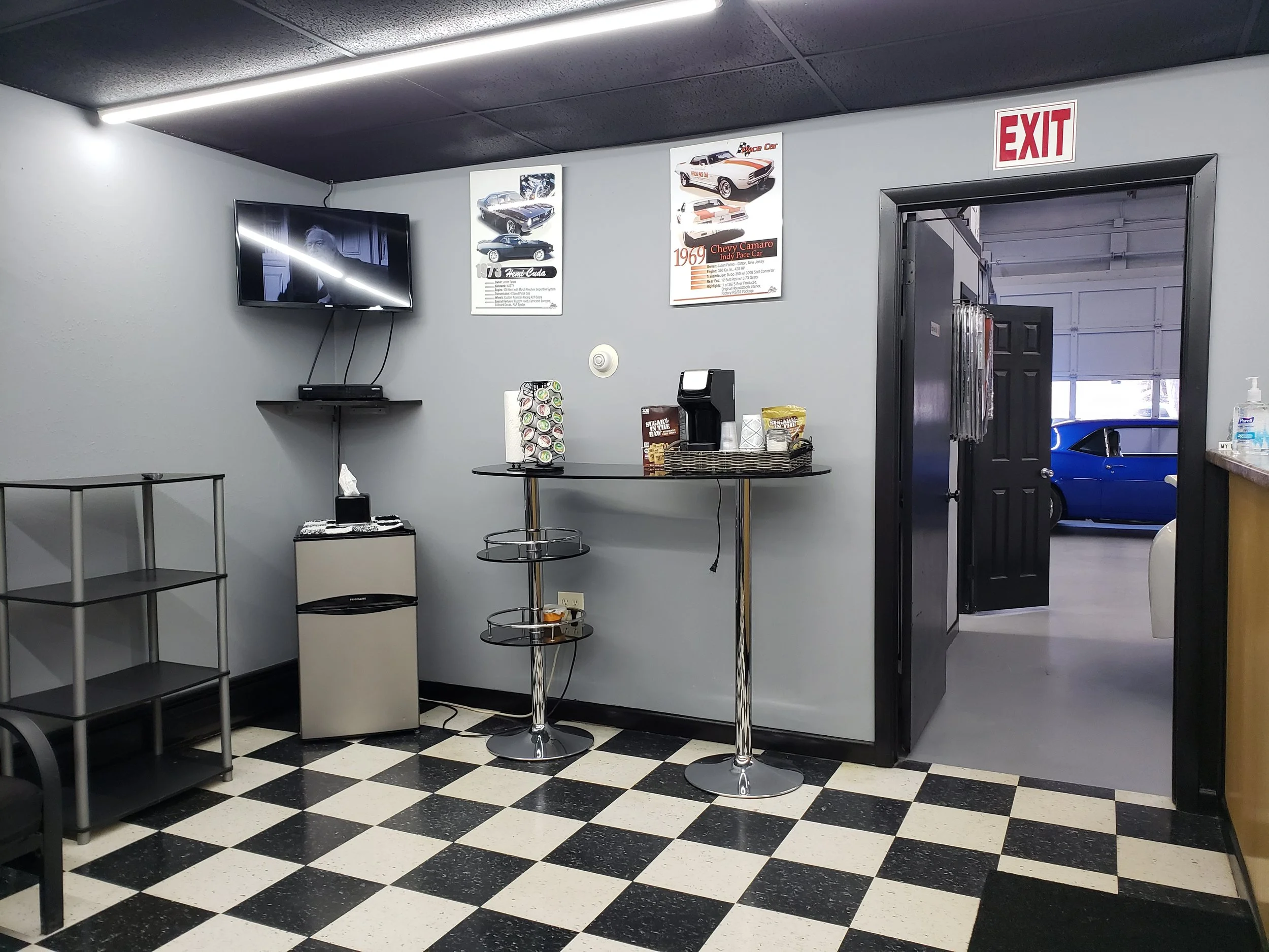 Interior of an automotive shop with a black and white checkered floor, a wall-mounted television, car posters, a black door leading to a garage, and a small refreshment area with a coffee machine and snacks at Towne Auto Repair in Randolph, NJ