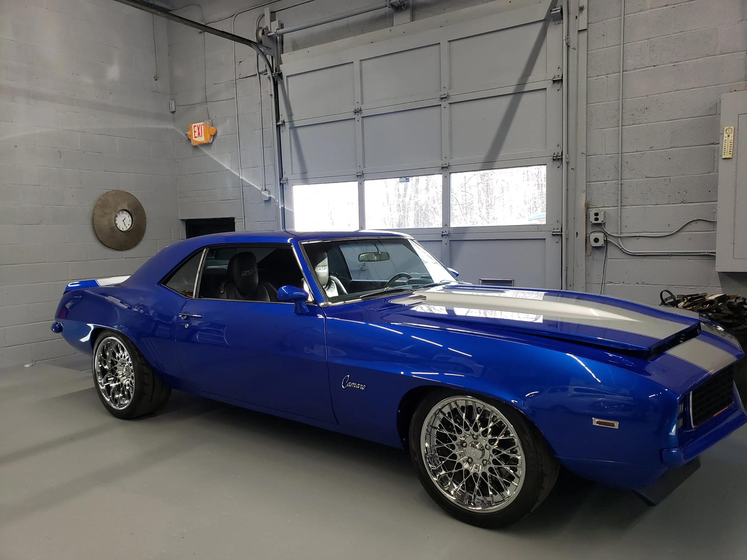A blue classic Chevrolet Camaro with chrome wheels parked inside a garage with gray walls and a large garage door at Towne Auto Repair, formerly Pete's Auto Repair in Randolph, NJ.