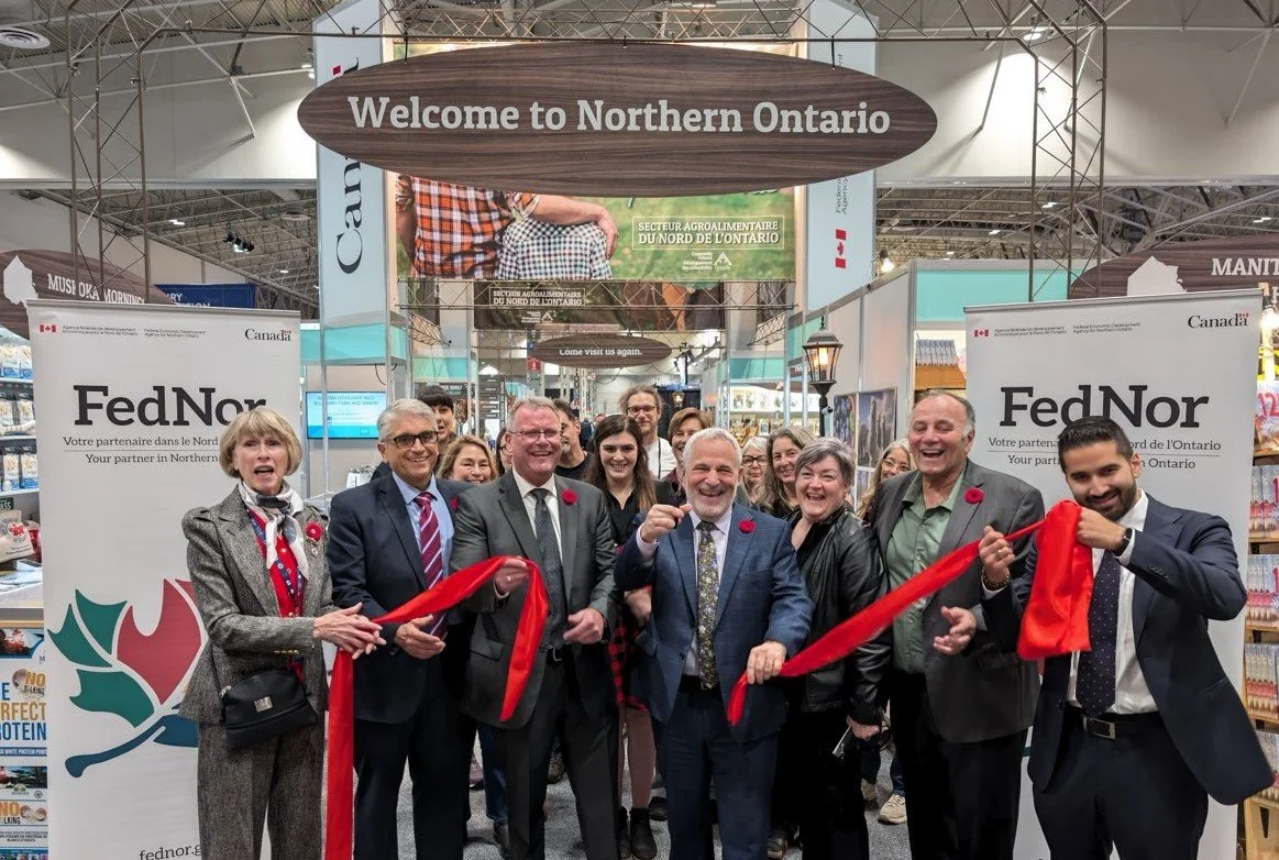 Group of people at the opening of a Northern Ontario booth, celebrating with a ribbon-cutting ceremony under a sign that reads 'Welcome to Northern Ontario' at an indoor exhibition or trade show.