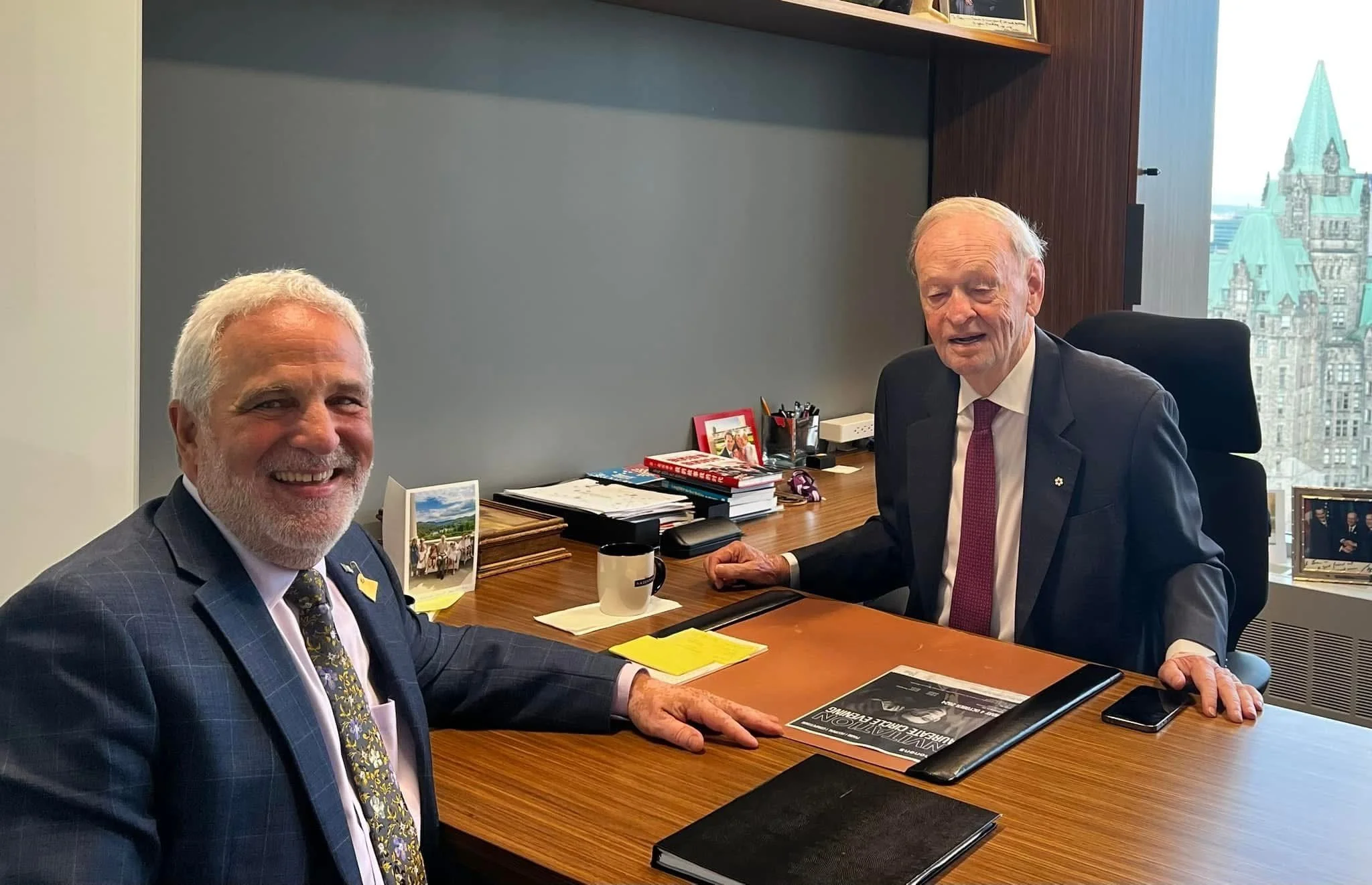 Two older men in suits sitting at a wooden desk in an office, smiling. One man has a beard and is smiling broadly, the other man has white hair and is looking slightly down. Office items and photographs are on the desk, with large windows showing a cityscape in the background.