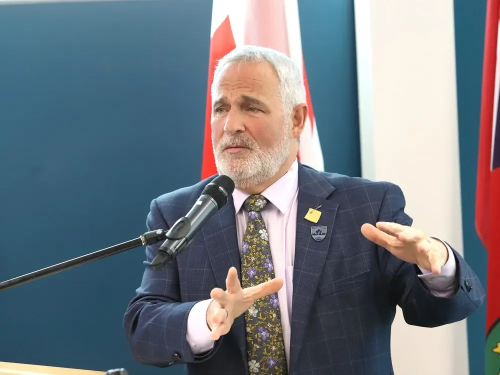 A man with gray hair and beard speaking at a podium with a microphone, gesturing with his hands, wearing a dark plaid suit and a colorful floral tie, with flags in the background.