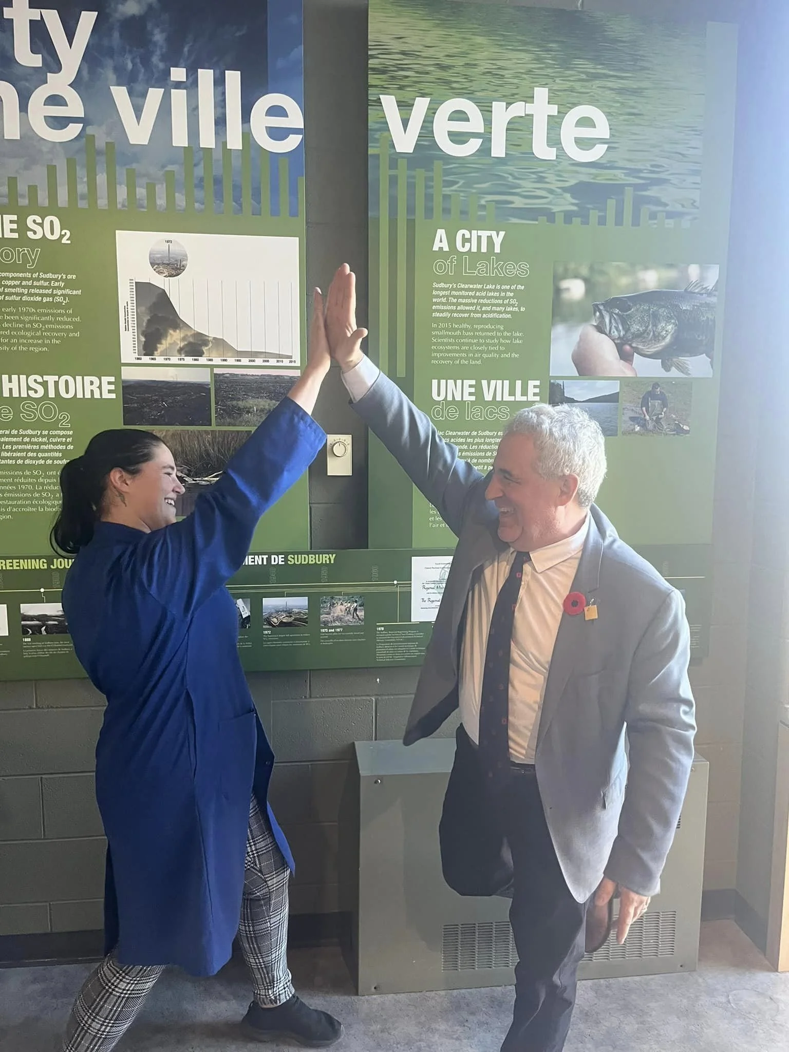 Two people high-fiving in front of an informational exhibit about the city of Lakes, with one person in a blue coat and the other in a gray blazer, both smiling.