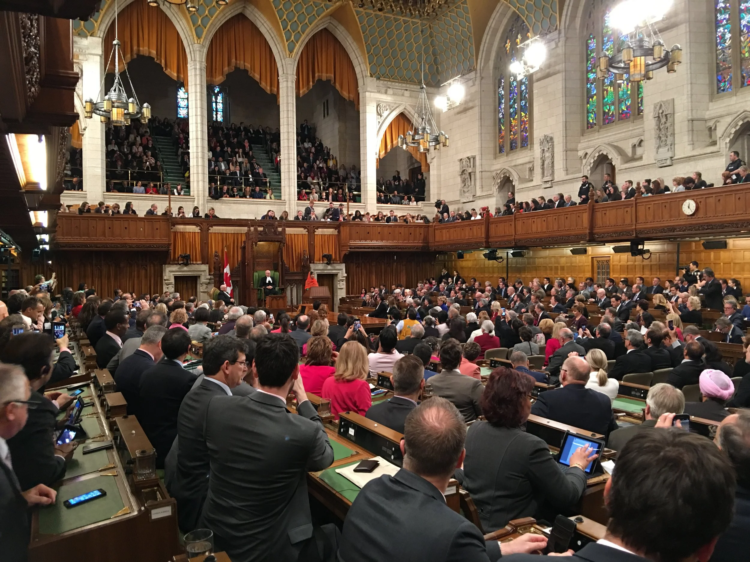 A government assembly in a large, ornate hall with wooden paneling, stained glass windows, and chandeliers. Many people are seated, some using devices, and speakers are standing at a podium.