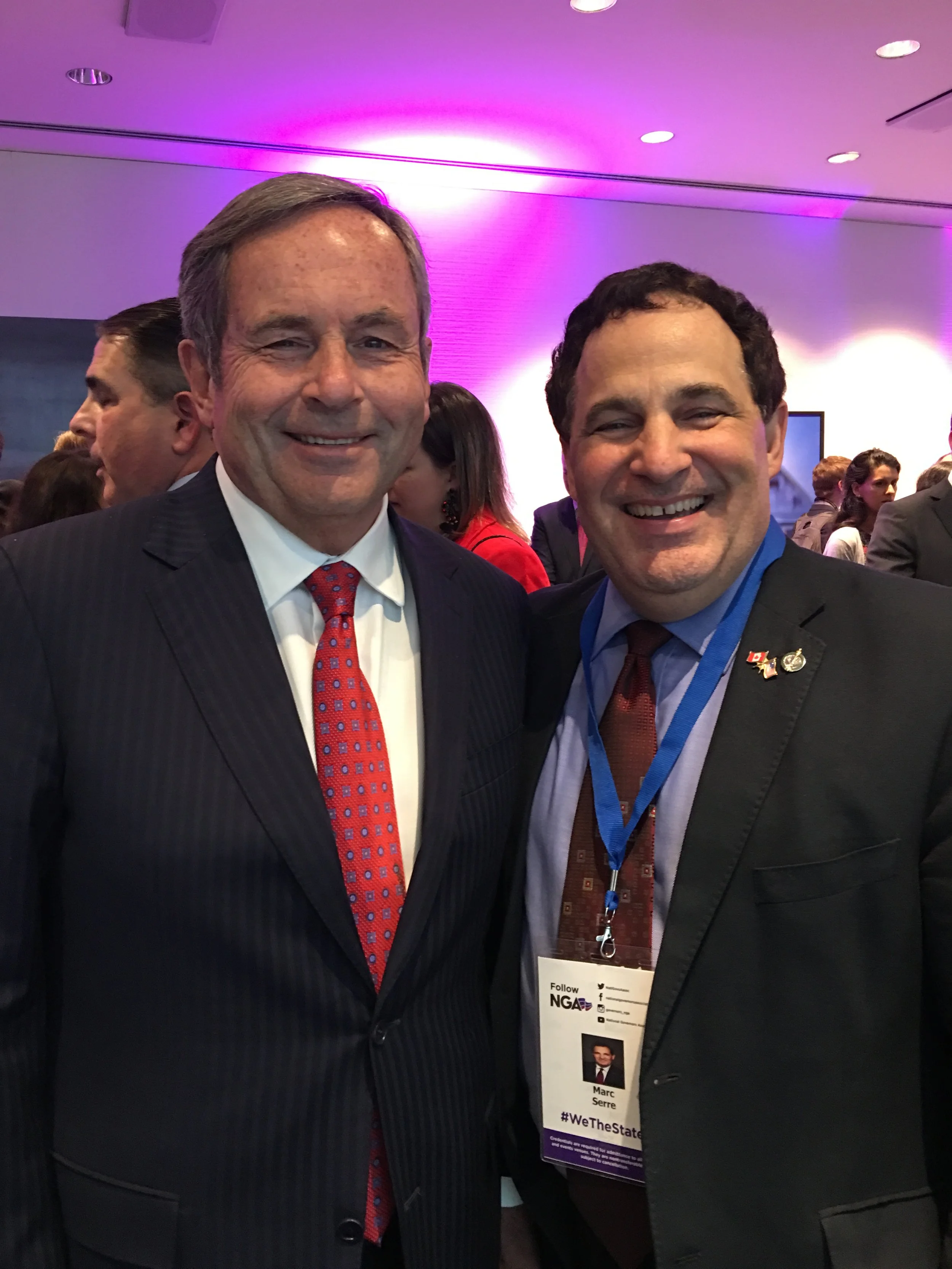 Two men in business attire at a conference, smiling for the camera, with a crowd in the background and pink lighting.