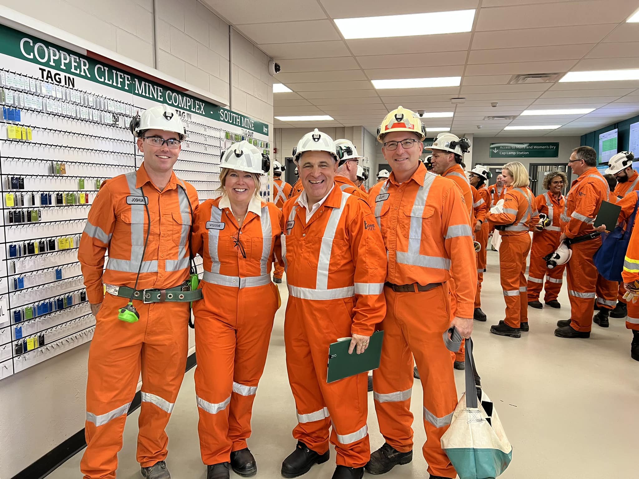 Group of workers in orange safety uniforms and helmets inside a mining facility, smiling at the camera, with a display of mining tags on the wall behind them.