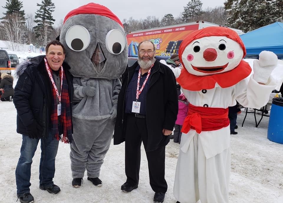Three people posing outdoors in winter, with two people in mascot costumes of a bird and a clown. Background includes snow, trees, and a colorful event tent.
