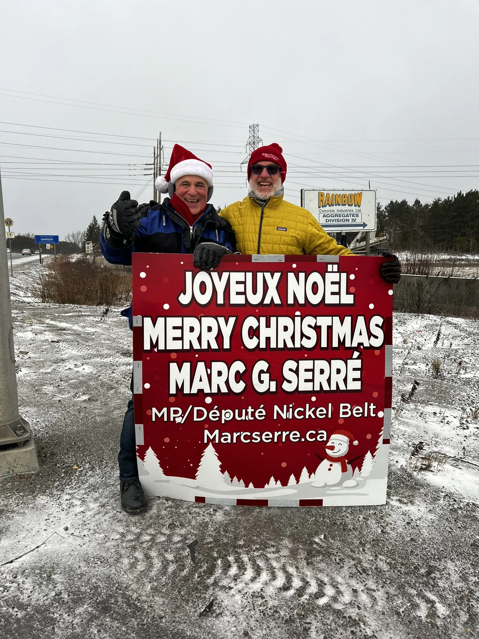 Two men are standing outdoors in winter, holding a Christmas sign that says "Joyeux Noël, Merry Christmas, Marc G. Serré" with snowman and winter trees illustrations, wearing winter jackets, hats, and gloves, with overcast sky and some snow on the gr