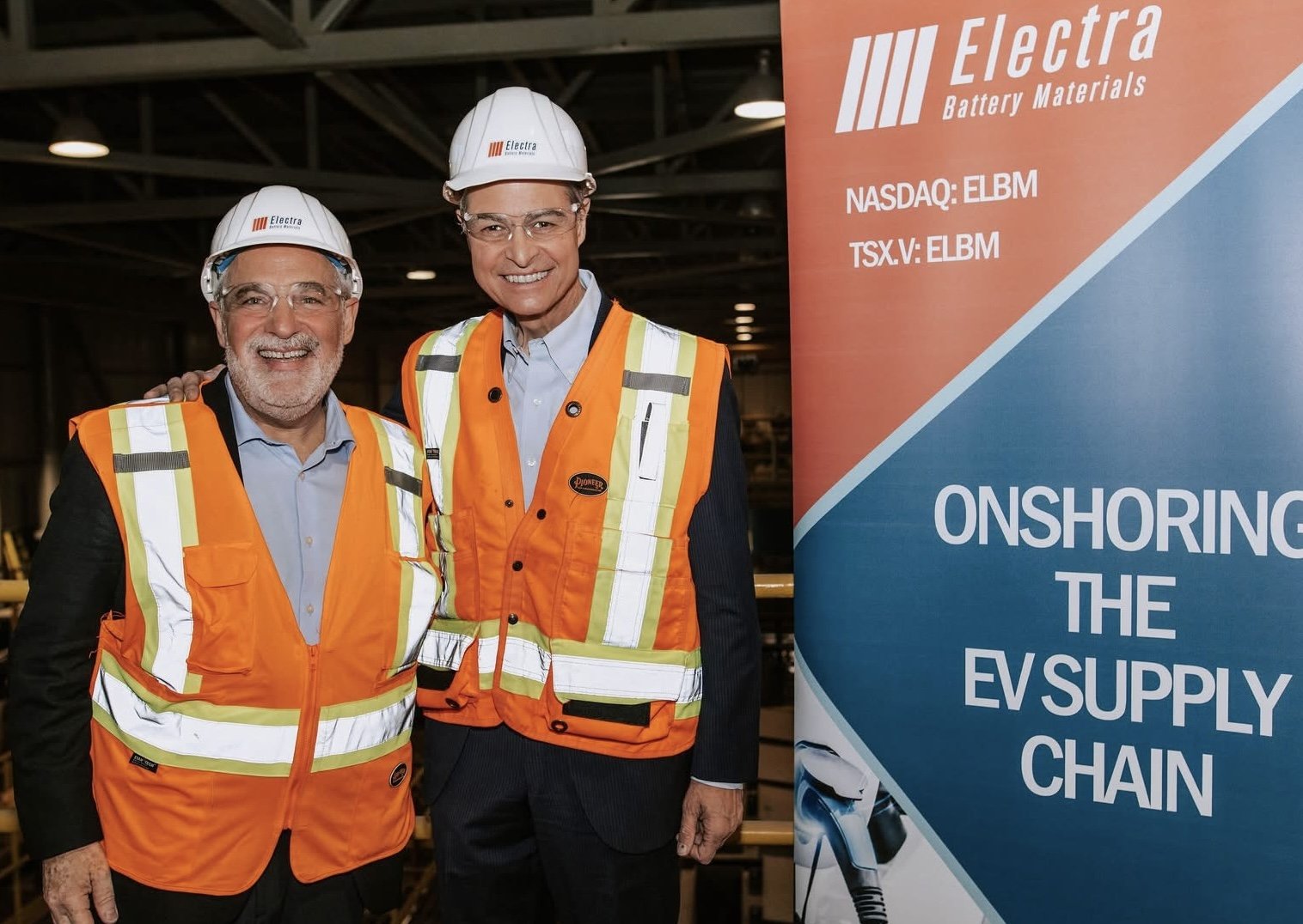 Two smiling men wearing white hard hats and orange safety vests stand together in an industrial setting. A large sign to their right reads 'Electra Battery Materials' and promotes onshoring the EV supply chain, with stock tickers NASDAQ:ELBM and TSX.