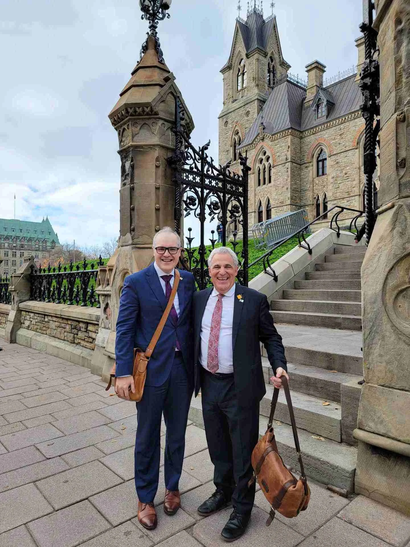 Two men in suits standing together outside in front of a historic building with gothic architecture, iron gates, and stone steps. They are smiling and appear happy.