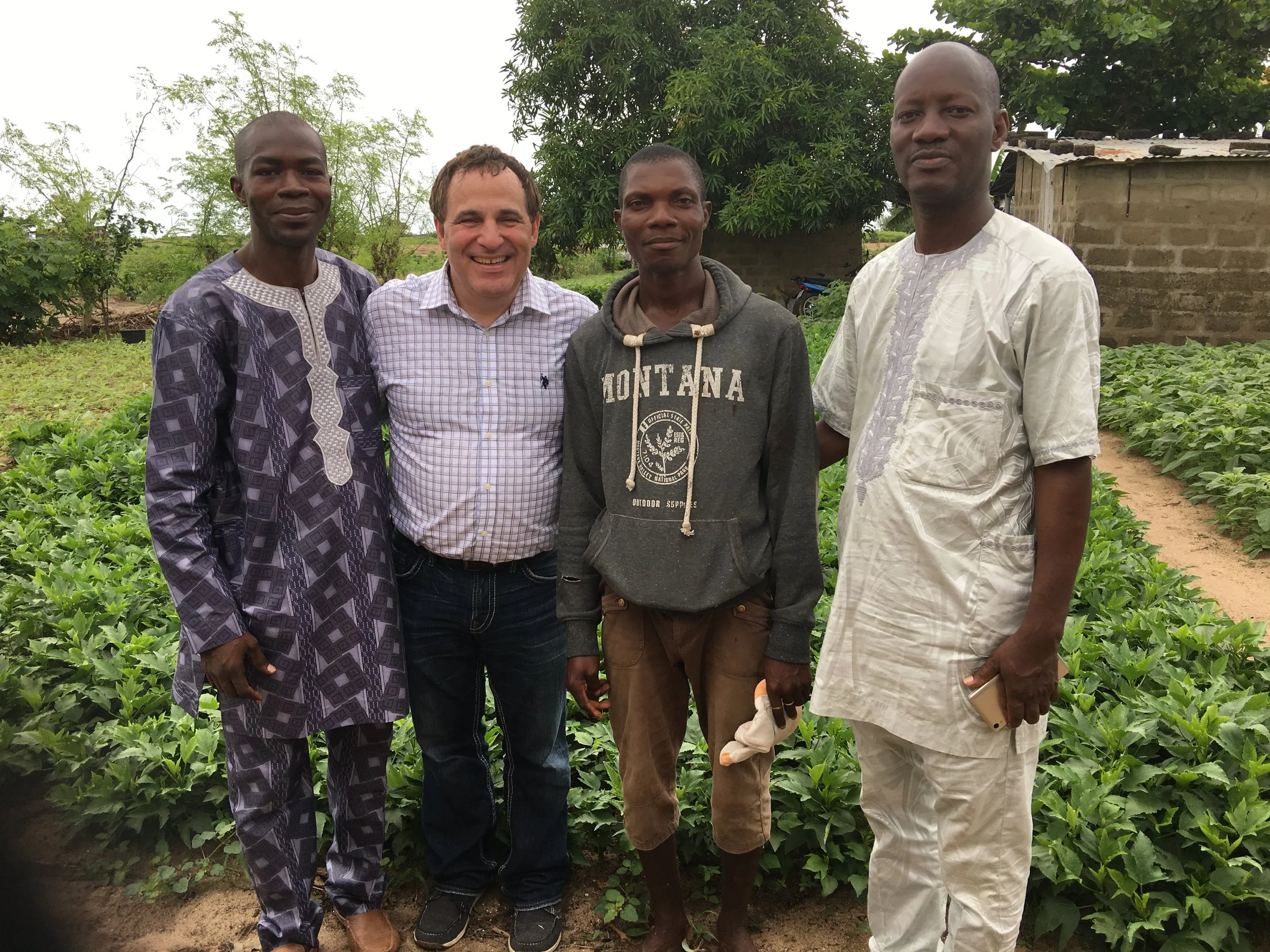 Five men standing together in a garden with green plants and trees, some holding mobile phones, smiling at the camera.