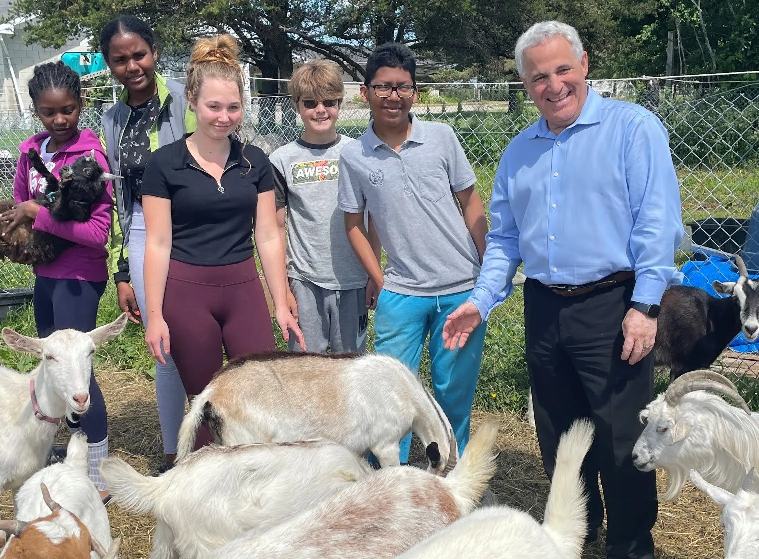 Group of people, including adults and children, standing outdoors with goats, a dog, and chickens in a fenced farm area.