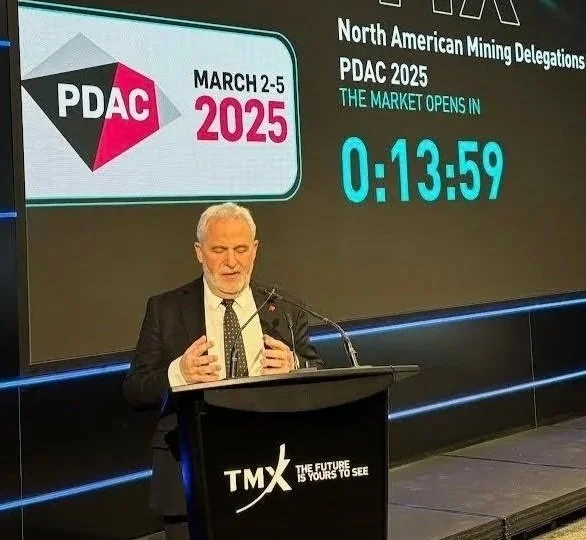 A man in a suit speaking at a podium with a large screen behind him displaying the text "North American Mining Delegations PDAC 2025" and a countdown timer. The event is scheduled for March 2-5, 2025, and the market opens in 13 minutes and 59 seconds.