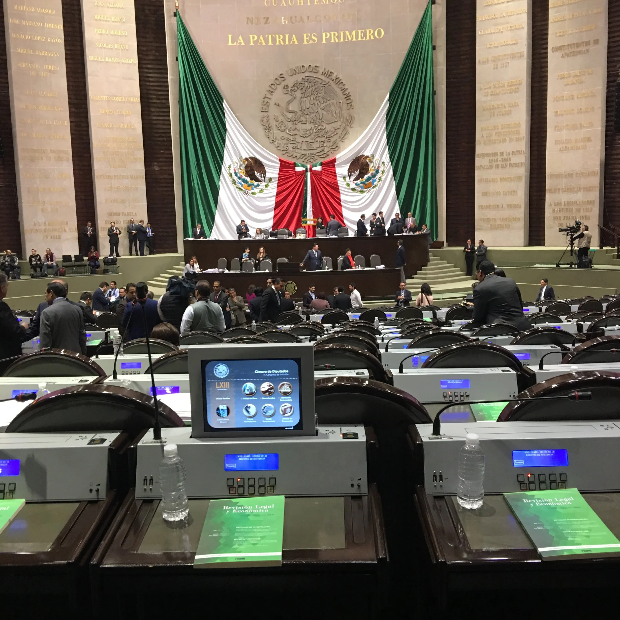View of the Mexican Congress chamber with a large Mexican flag and emblem behind the speaker's area, and politicians and officials preparing and conversing at their desks.