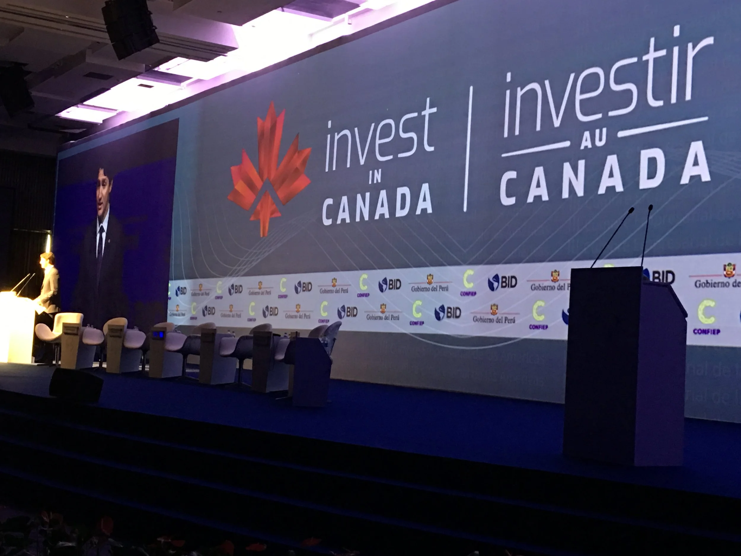 Conference stage with a large screen displaying the logos and texts 'Invest in Canada' and 'Invest au Canada.' A woman is speaking at a podium to the left, and there are empty chairs on the stage. The backdrop features logos of BID, Gobierno del Peru, and CONFEIP.