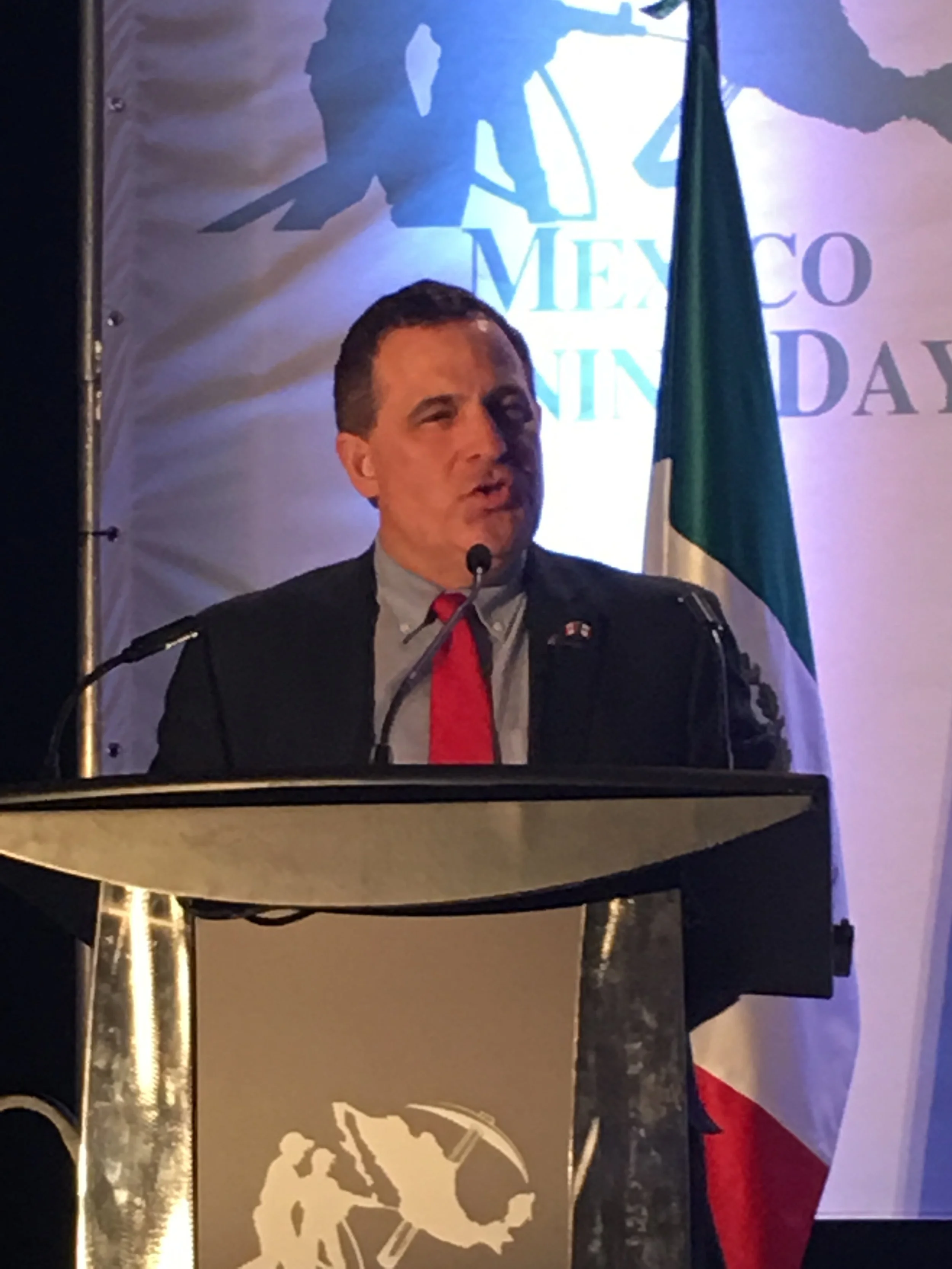 A man in a dark suit and a red tie speaking at a podium with a microphone, behind a Mexican flag, at an event celebrating Mexico Independence Day.