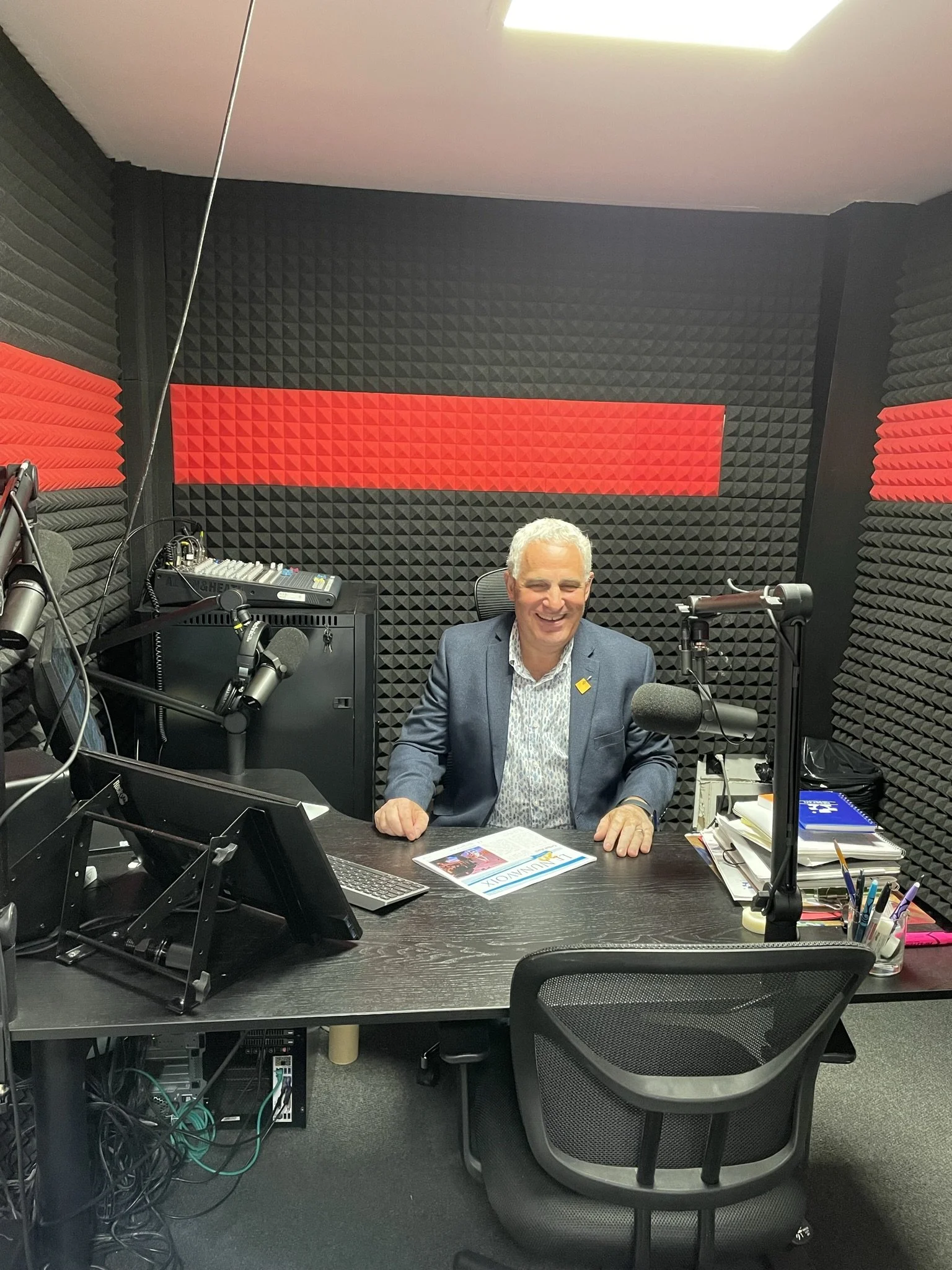 A man with gray hair dressed in a blue blazer and a light patterned shirt sitting in a professional radio studio with soundproof foam panels, microphones, and audio equipment, smiling at the camera.