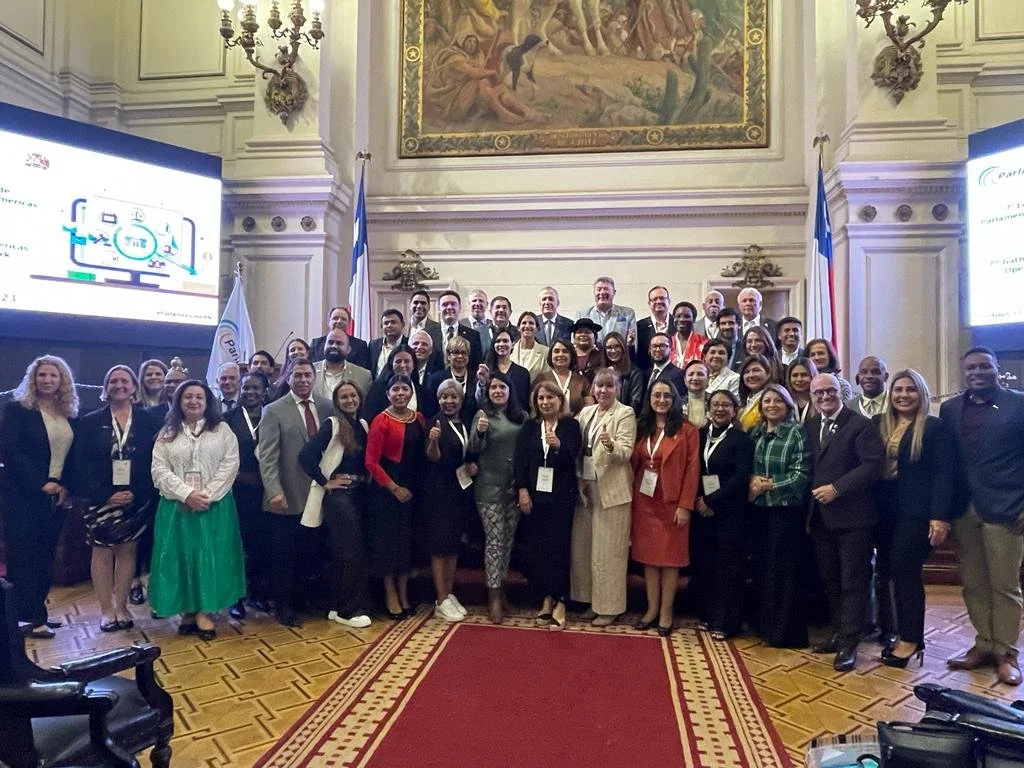 A large group of diverse people posing for a photo in an opulent, ornate room with high ceilings, a large painting, flags, and a projector screen. The group includes men and women dressed in business attire, standing on a carpeted floor with intricate wood flooring around it.