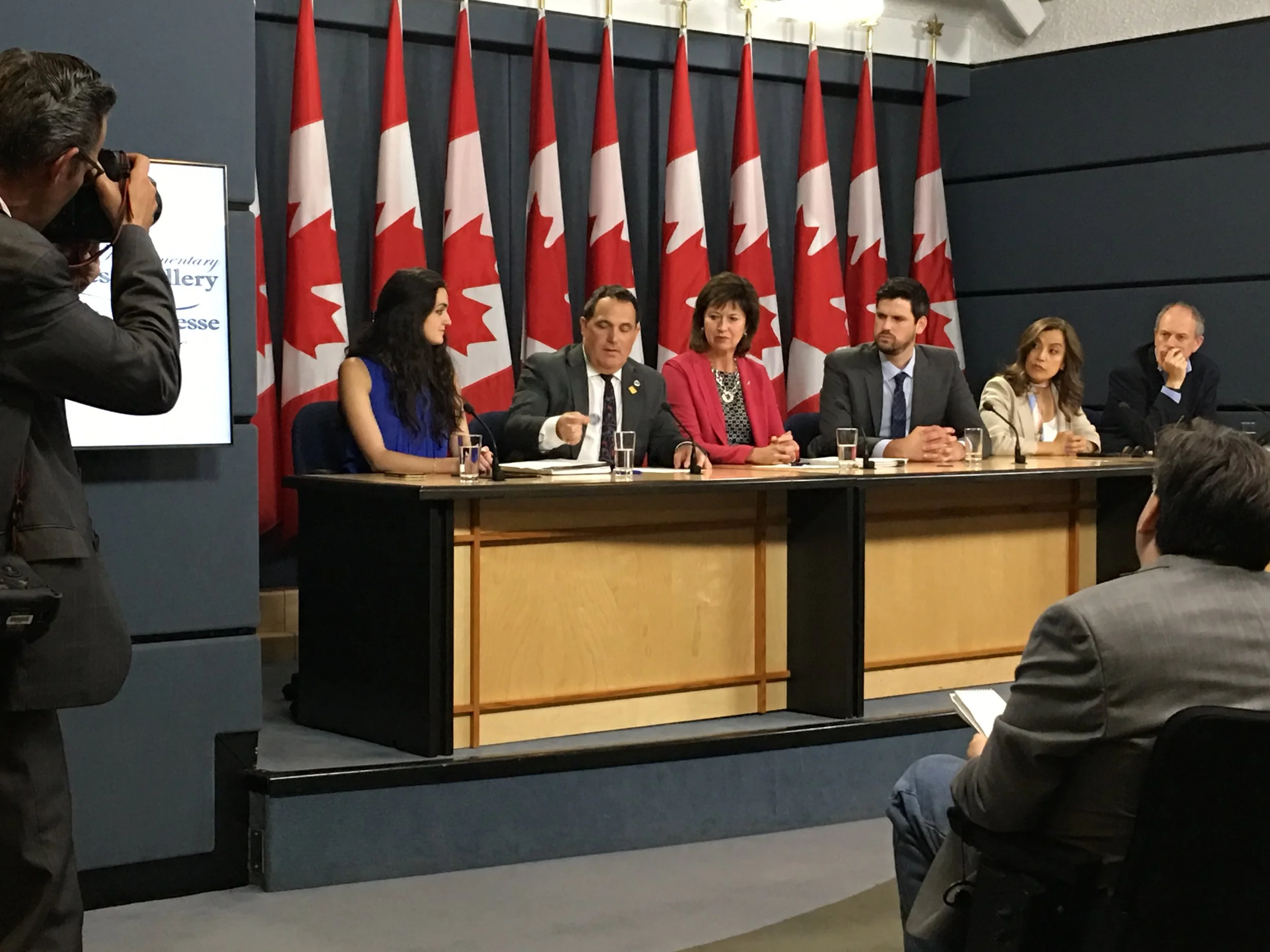 A panel of six people at a press conference, seated at a long table with microphones and glasses of water, behind a row of Canadian flags. A man in a suit is speaking, while a photographer takes photos from the left.