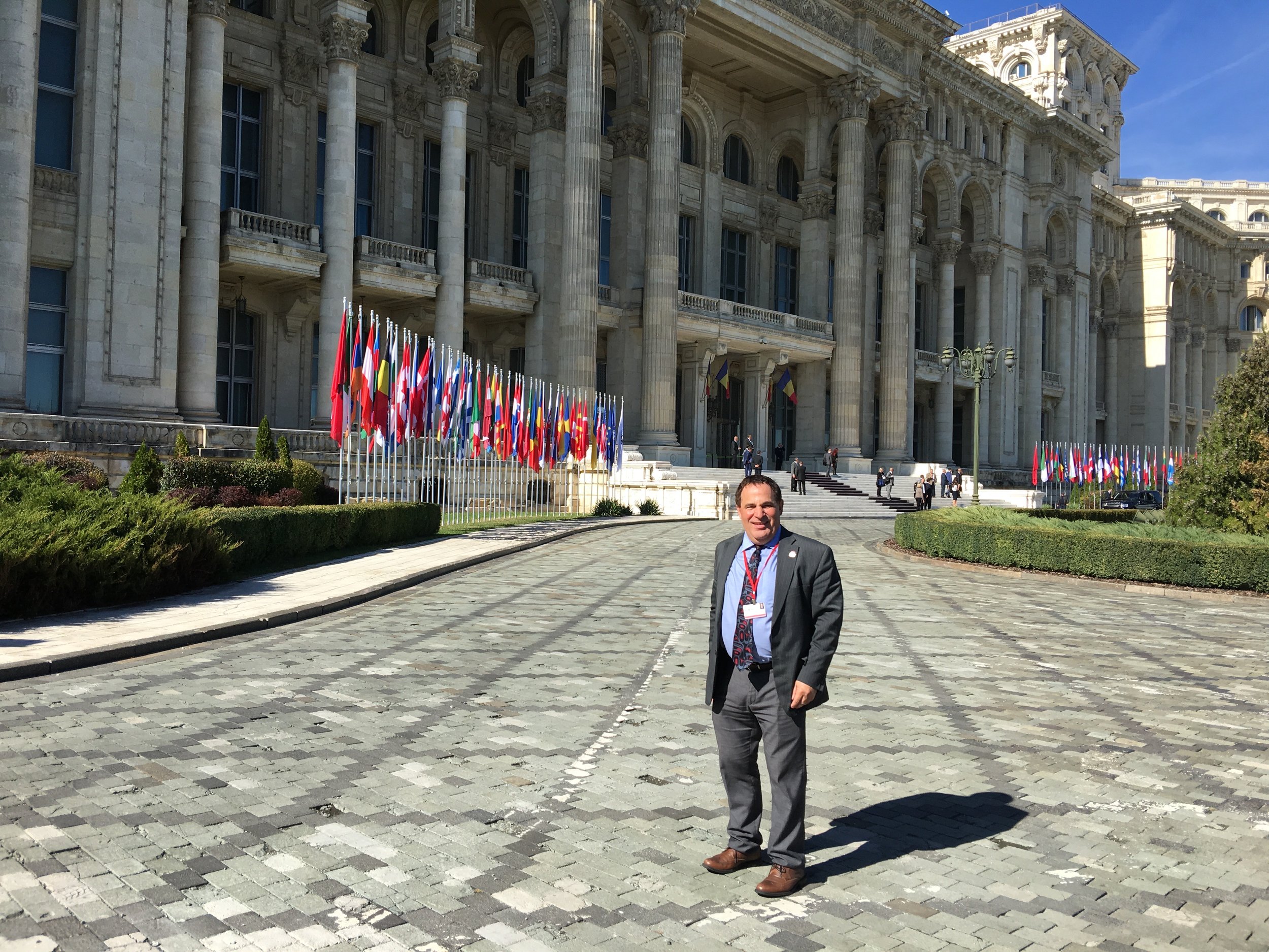 A man in a suit standing on a cobblestone street in front of a grand government building with flags from various countries displayed on flagpoles outside. The sky is clear and sunny.