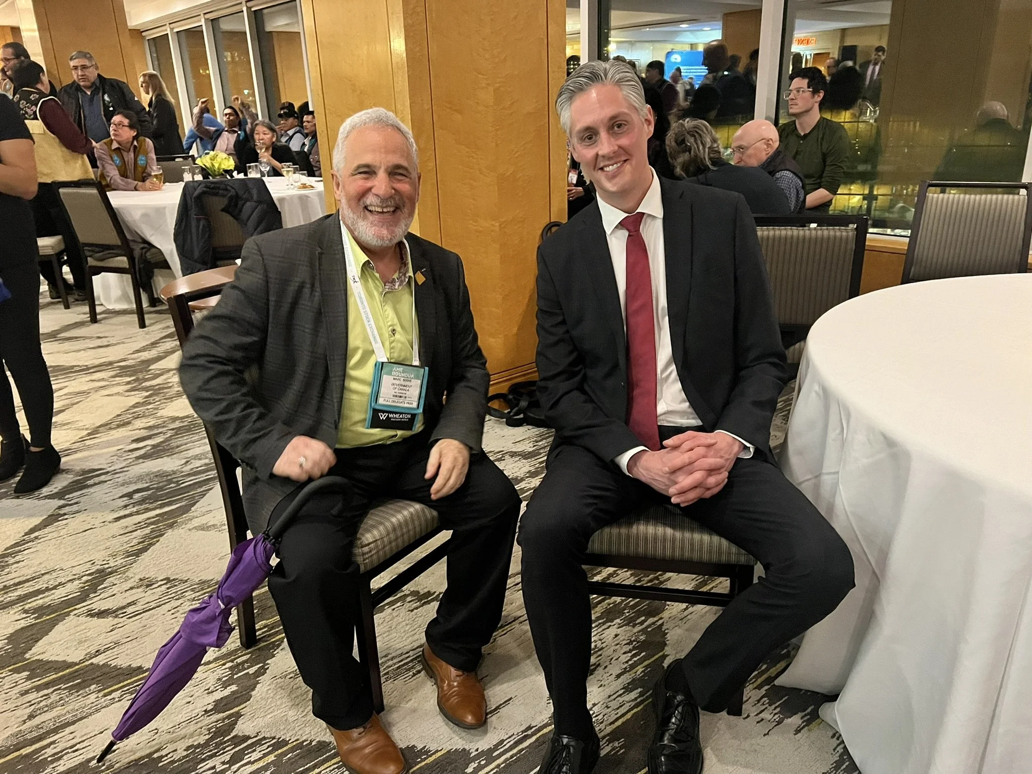 Two men in formal attire sitting at a banquet table, smiling for the camera, at a crowded indoor event.