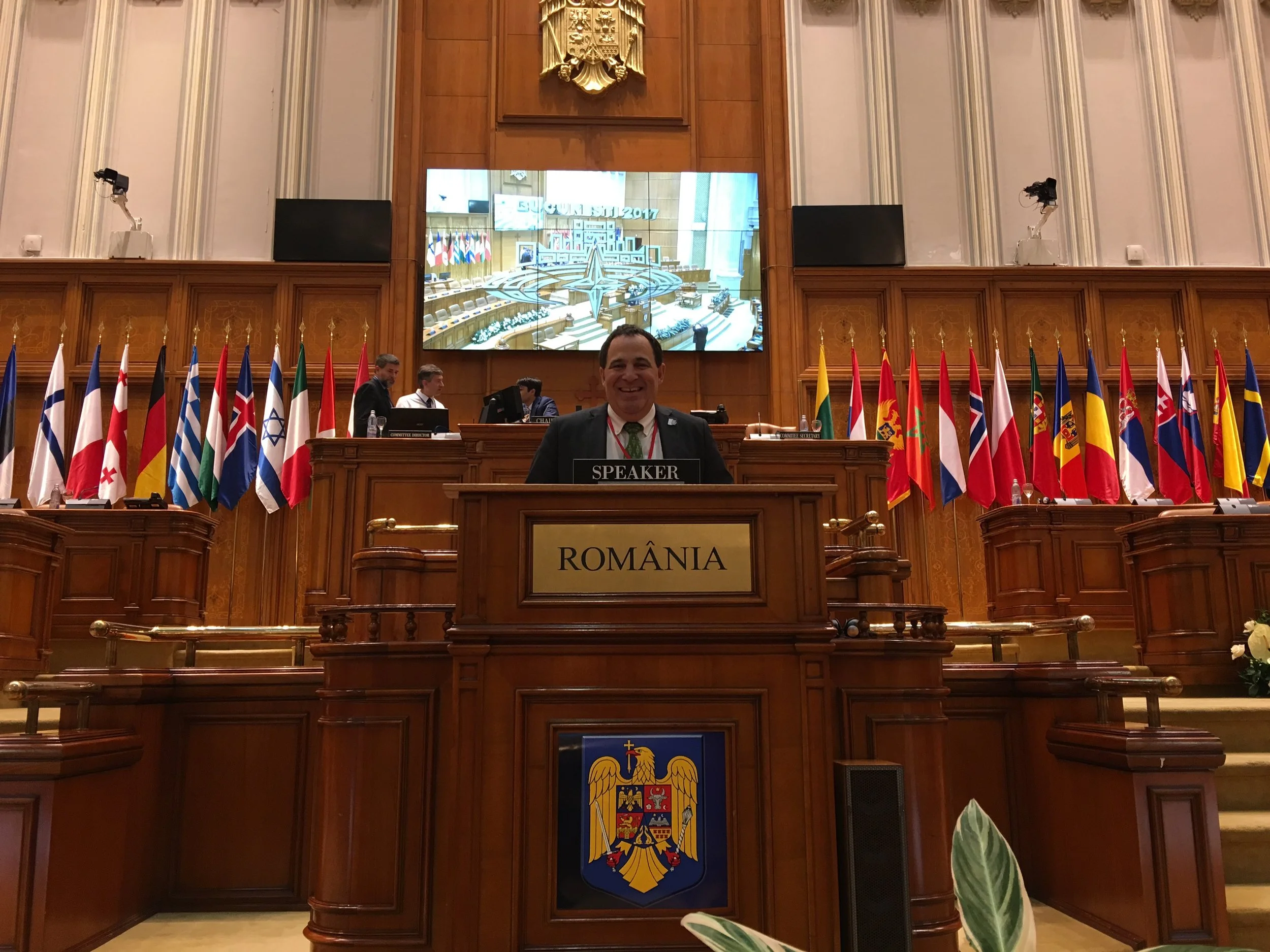 A man smiling at an official's desk labeled 'ROMANIA' and 'SPEAKER' in a room with many national flags, with a large screen behind showing a conference room with flags and a screen.